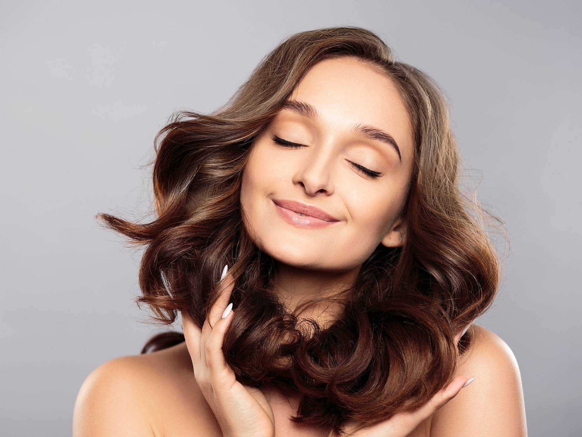 Frau mit langen, lockigen braunen Haaren, die ihre Haare mit beiden Händen hält, vor grauem Hintergrund