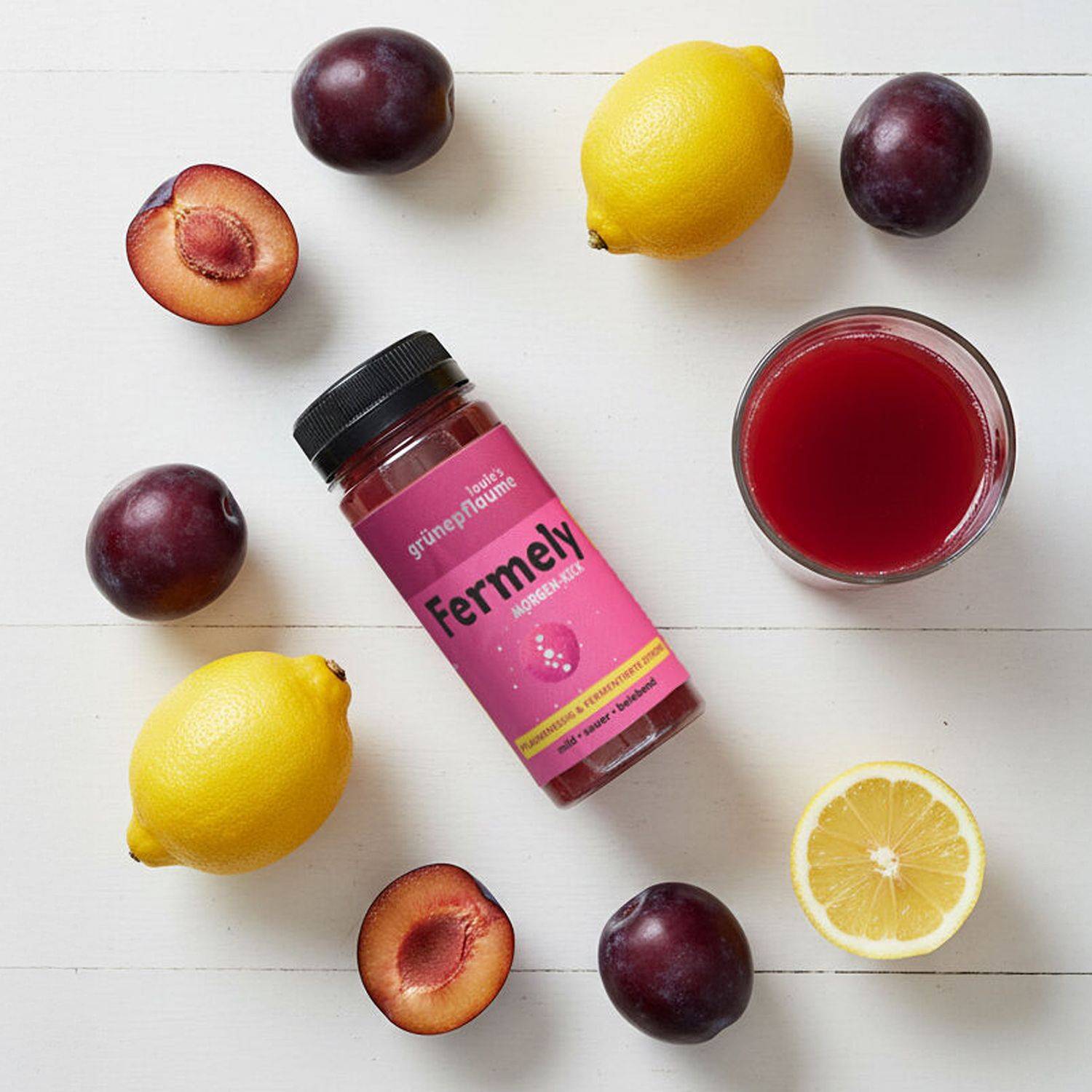 Flasche mit rotem Getränk 'Fermely' von 'louie's grüne pflaume' liegt auf weißer Fläche, umgeben von ganzen und halbierten Pflaumen und Zitronen, daneben ein Glas mit rotem Saft.