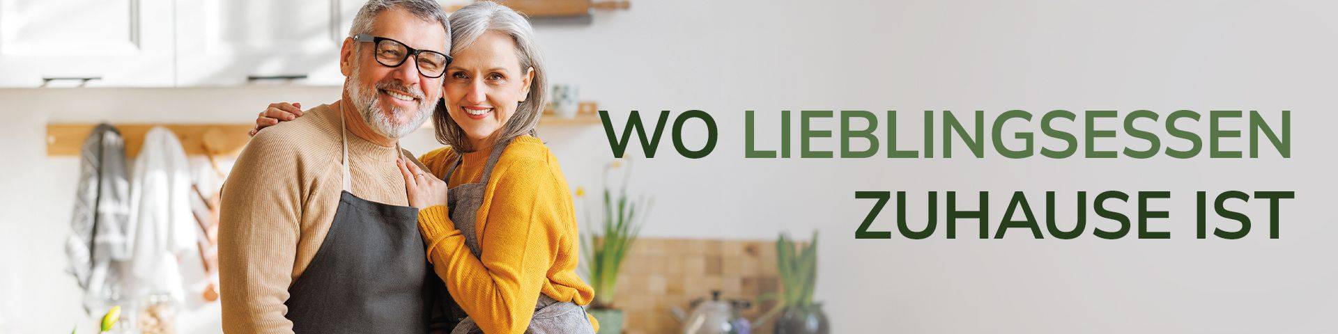 Älteres Paar umarmt sich in einer Küche, beide tragen Schürzen, Text 'WO LIEBLINGSSESSEN ZUHAUSE IST' in grüner Schrift rechts.