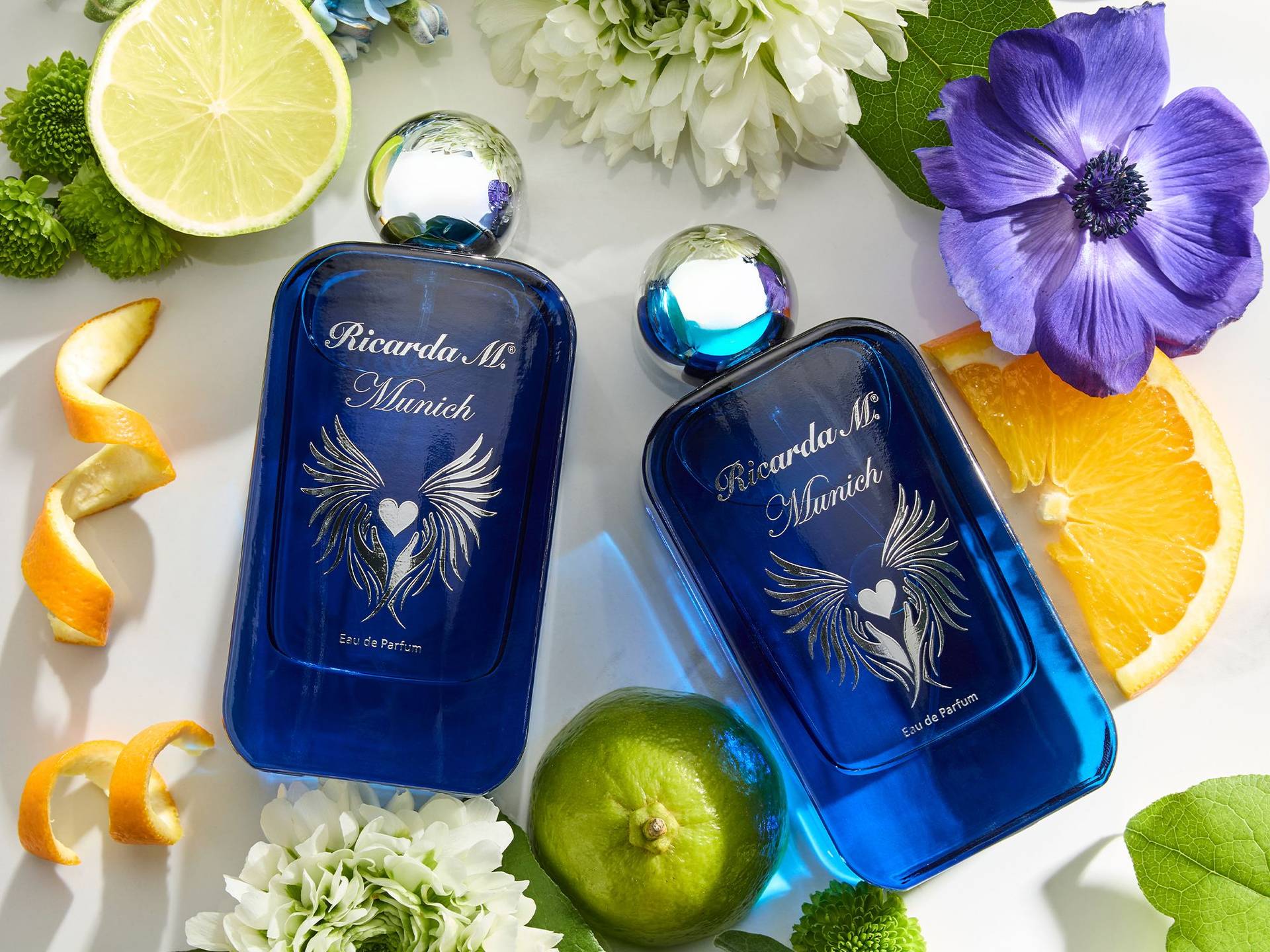 Zwei blaue Parfümflaschen mit silbernen Kugeldeckeln und der Aufschrift 'Ricarda M. Munich' umgeben von Limetten-, Orangen- und Zitronenscheiben sowie weißen und violetten Blüten.