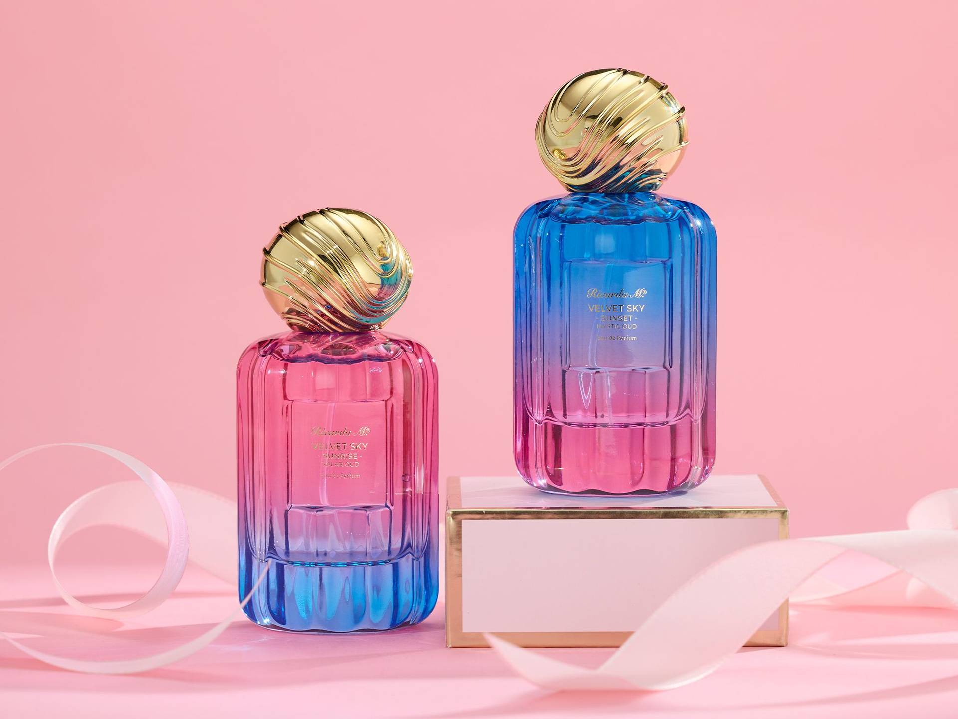 Zwei Flakons des Parfums Velvet Sky mit goldfarbenen, kugelförmigen Verschlüssen vor rosa Hintergrund, einer steht auf einem weißen Podest mit goldener Kante