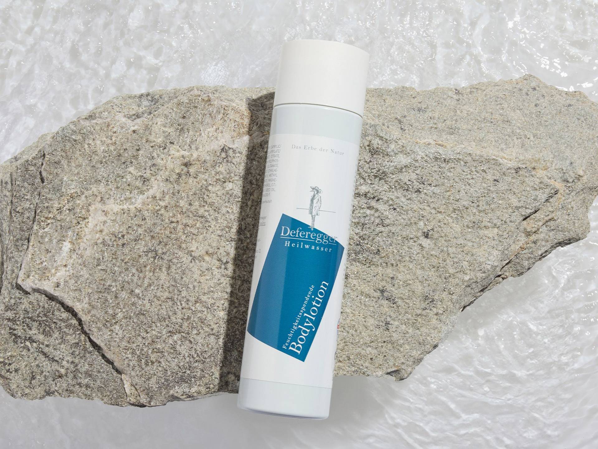 Weiße Flasche mit blauem Etikett und Aufschrift "Deferegger Heilwasser Bodylotion" liegt auf zwei großen grauen Steinen