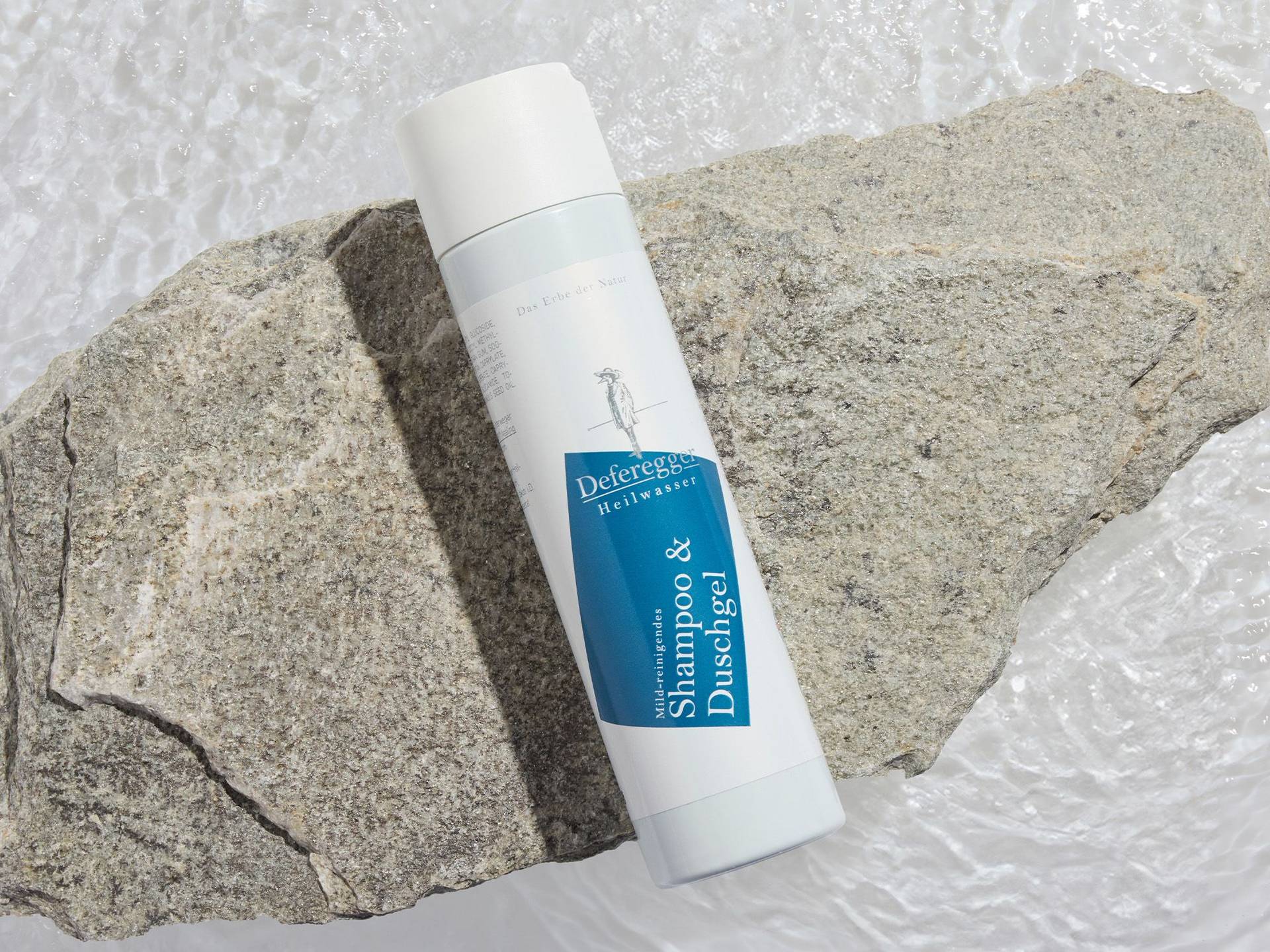 Weiße Flasche mit blauem Etikett und der Aufschrift 'Deferegger Heilwasser Shampoo & Duschgel' liegt auf zwei grauen Steinen