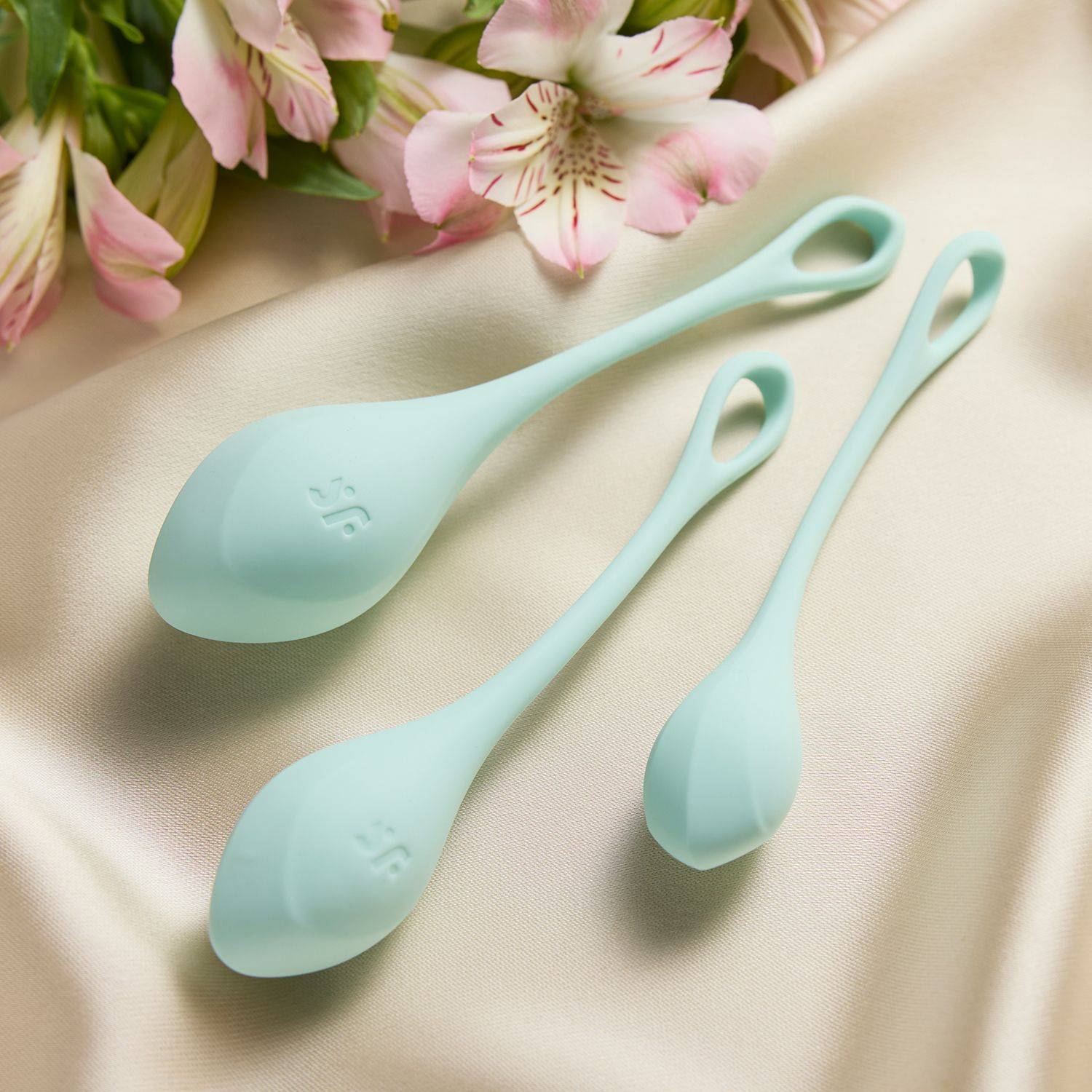 Drei unterschiedlich große, hellblaue Silikon-Liebeskugeln mit Schlaufe auf cremefarbenem Stoff und rosa Blüten 