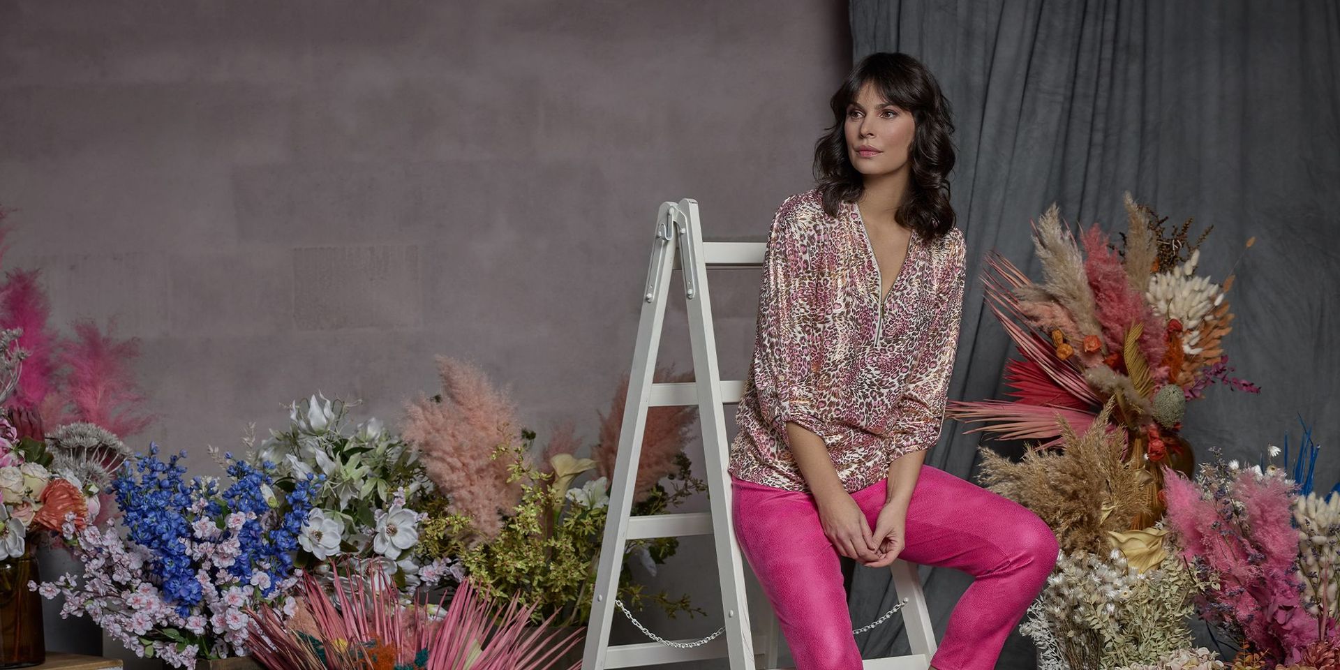 Dunkelhaarige Frau auf Leiter sitzend, um sie herum ganz viele Blumen.