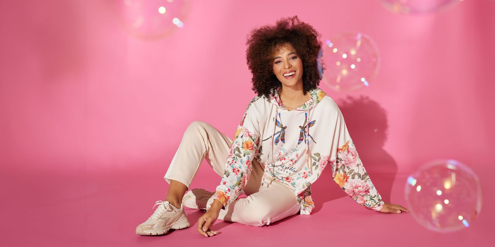 Frau in floralem Hoodie und beigen Hosen vor rosa Hintergrund, Mode von CHANNEL21
