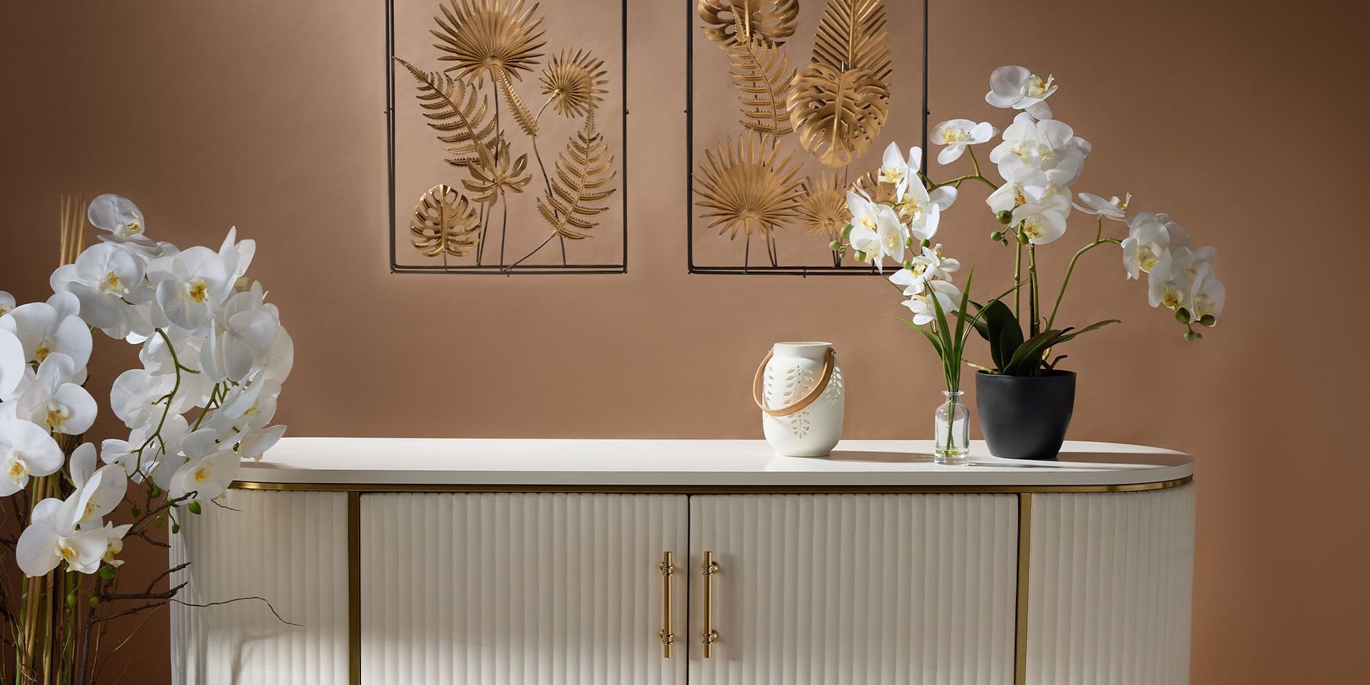 Orchideen und Windlicht stehen auf einer Kommode. An der Wand hängt eine Blumen-Wanddekoration