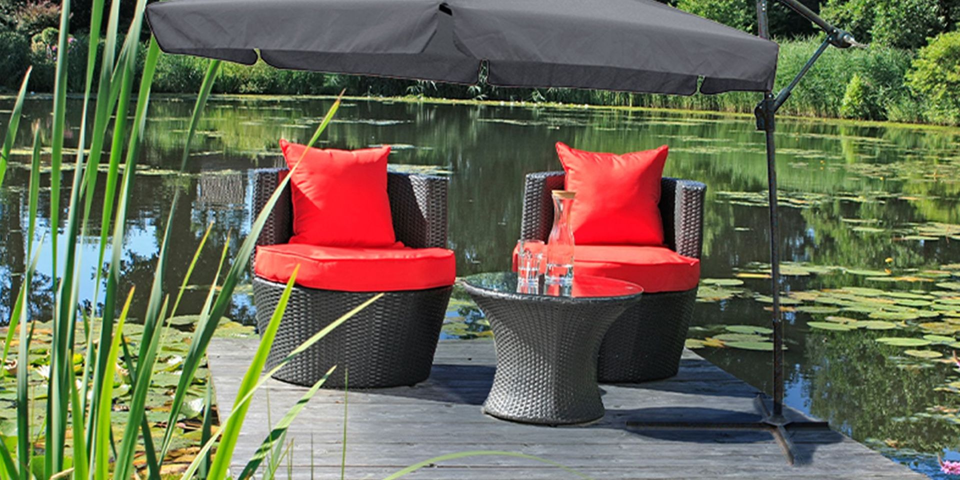 Zwei Rattan-Sessel mit roten Kissen und Glastisch auf Holzterrasse am See