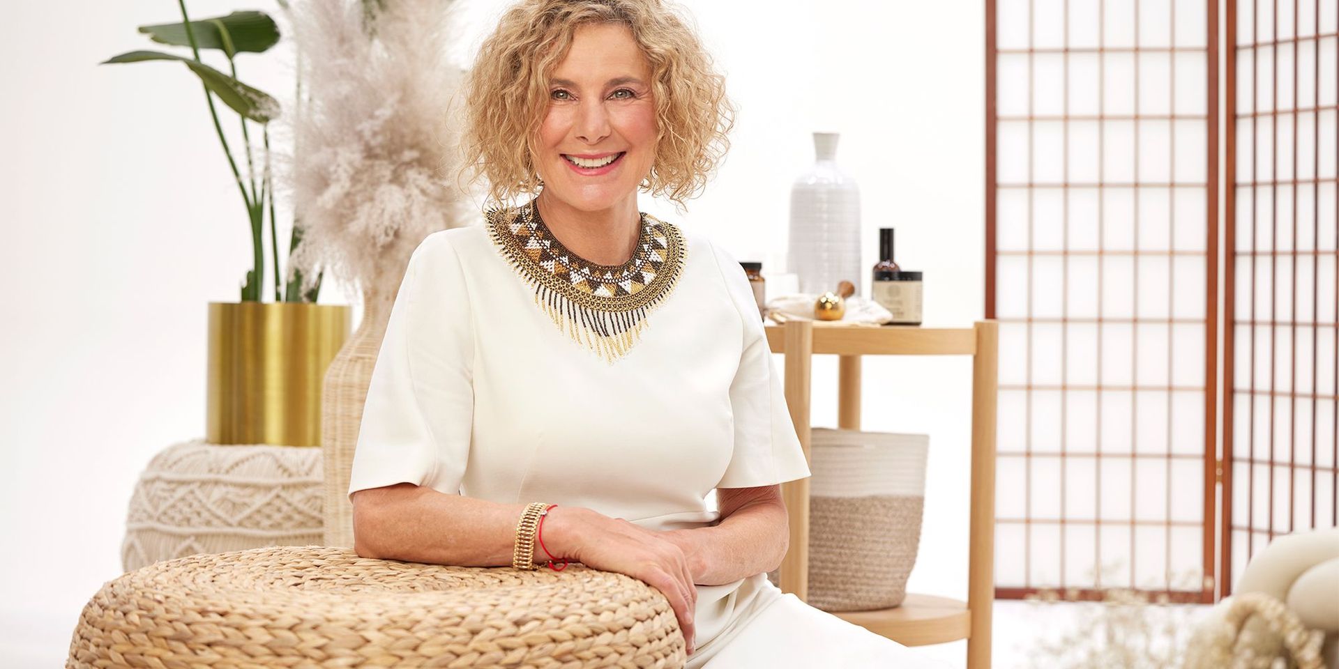 Frau in weißem Kleid mit auffälliger Halskette, sitzt auf geflochtenen Körben, umgeben von dekorativen Vasen und Wellnessprodukten.