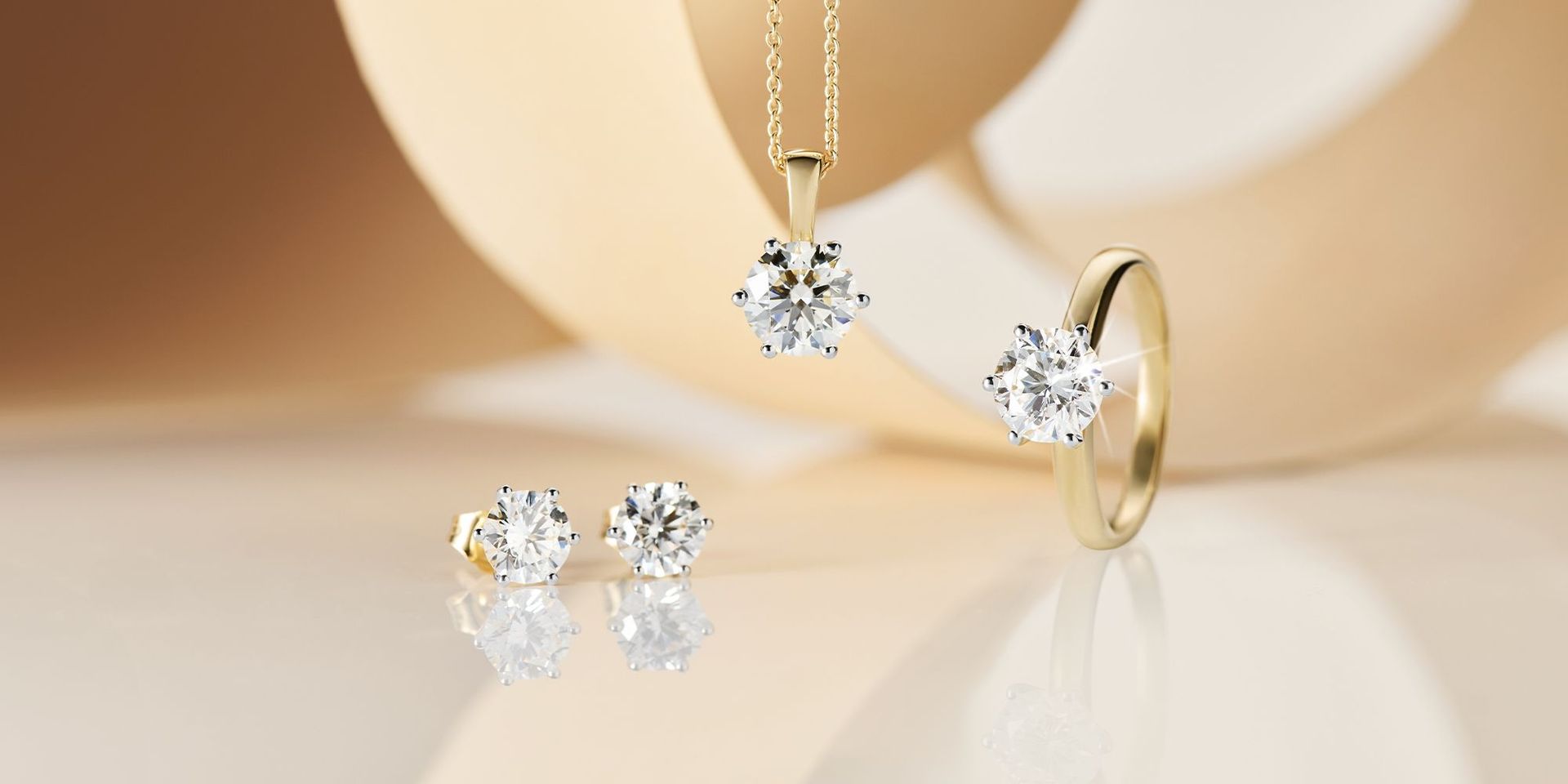 Diamantohrstecker, ein Diamant-Solitärring und ein Diamantanhänger vor einem hellgelben Hintergrund