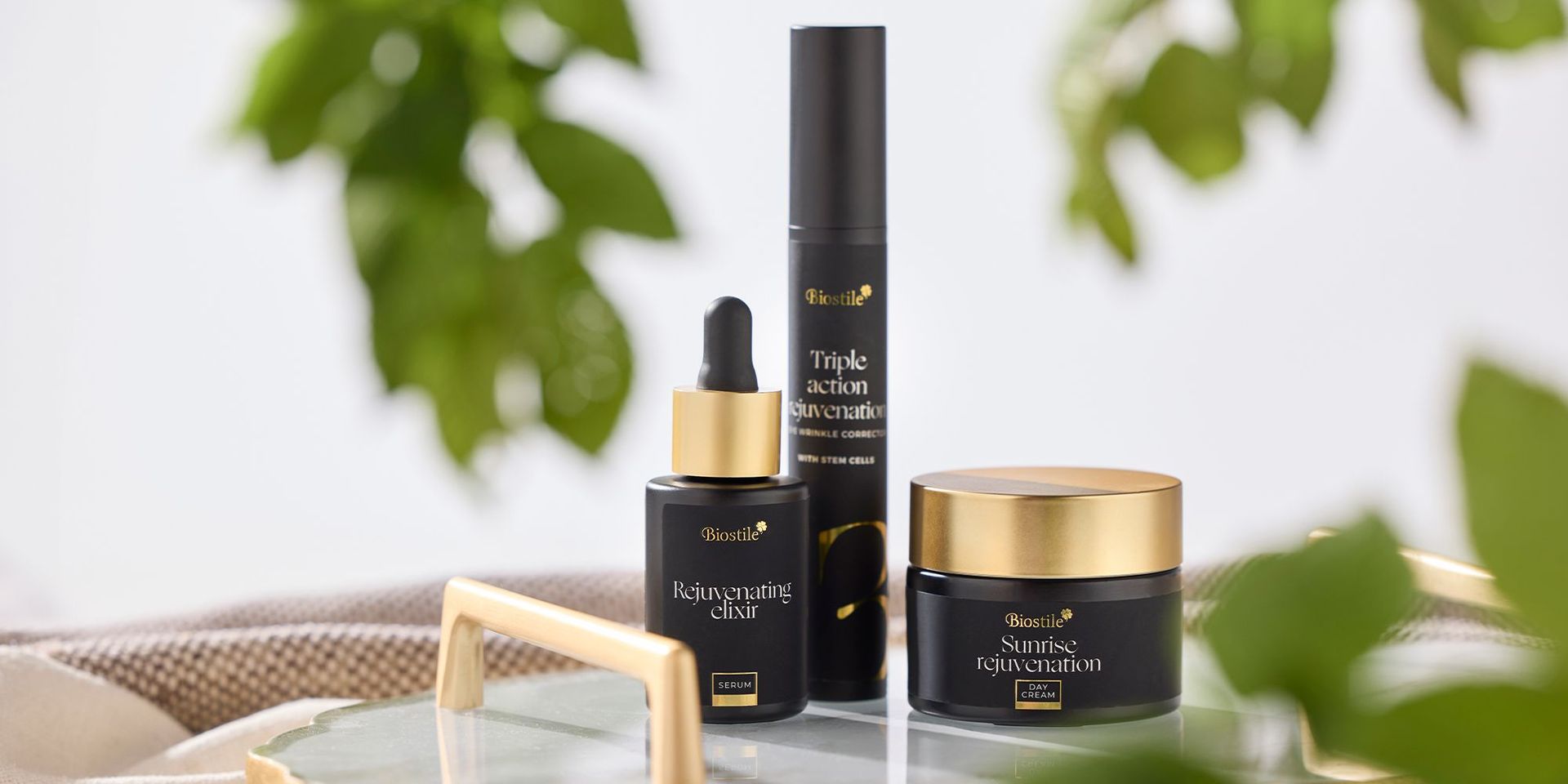 Drei schwarze und goldene Biostile Hautpflegeprodukte: Rejuvenating Elixir Serum, Triple Action Rejuvenation und Sunrise Rejuvenation Tagescreme auf Glasfläche