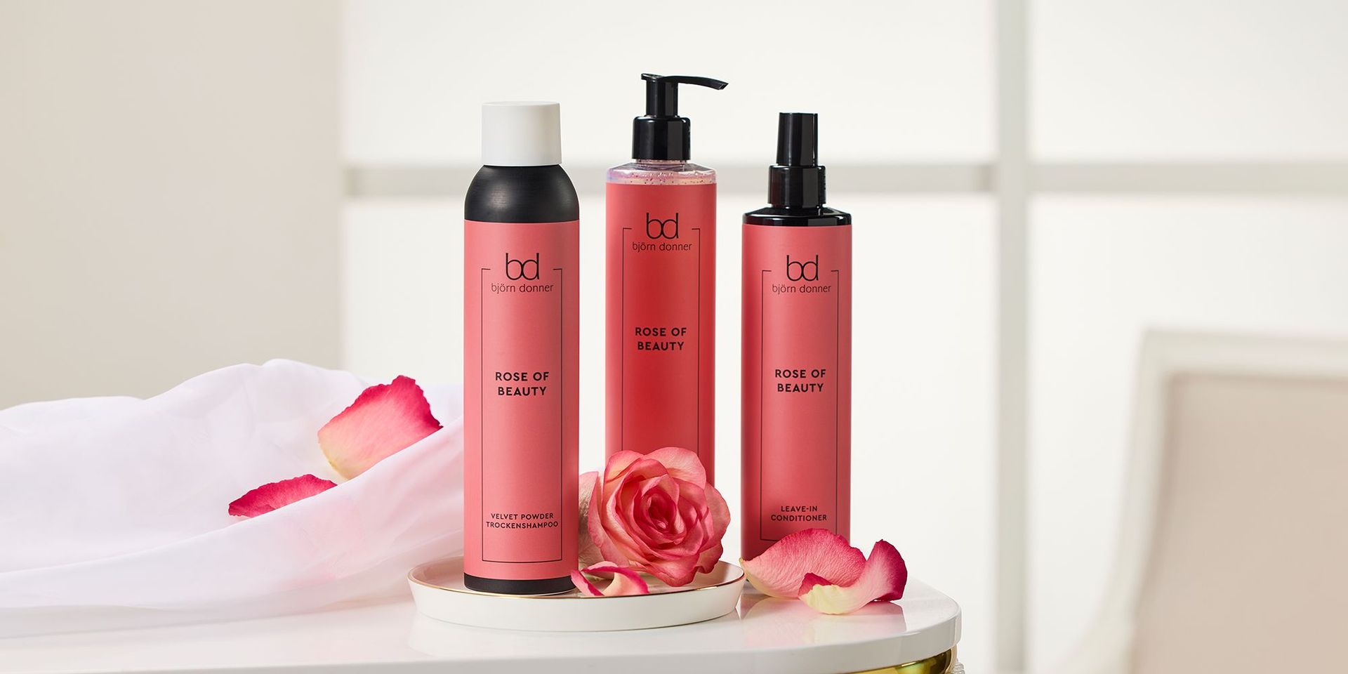 Drei rosafarbene Björn Donner Haarpflegeprodukte der Linie Rose of Beauty auf einem weißen Tisch, umgeben von Rosenblättern und einer Rose.