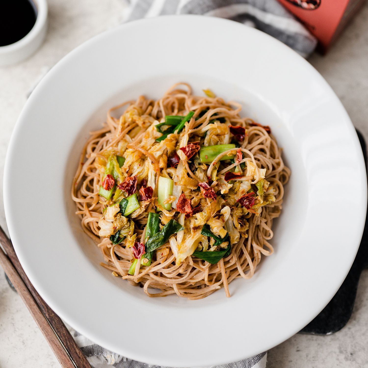 Teller mit gebratenen Nudeln, Gemüse und roten Chilistücken, daneben eine Packung Just Taste Pasta, Sojasauce und Knoblauch