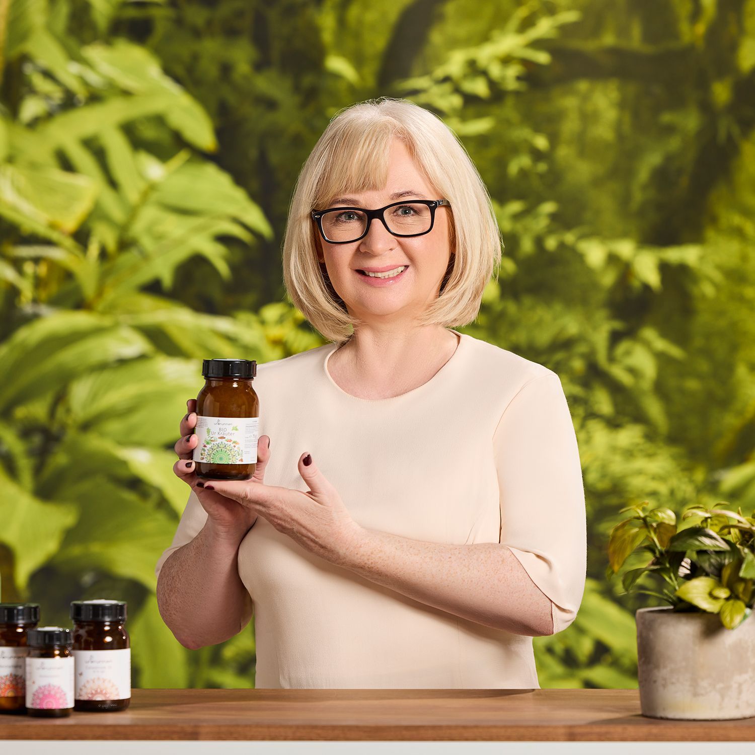 Urbrunnen-Expertin Monika Stedler hält braunes Glasfläschchen mit weißem Etikett, steht vor grünem Pflanzenhintergrund