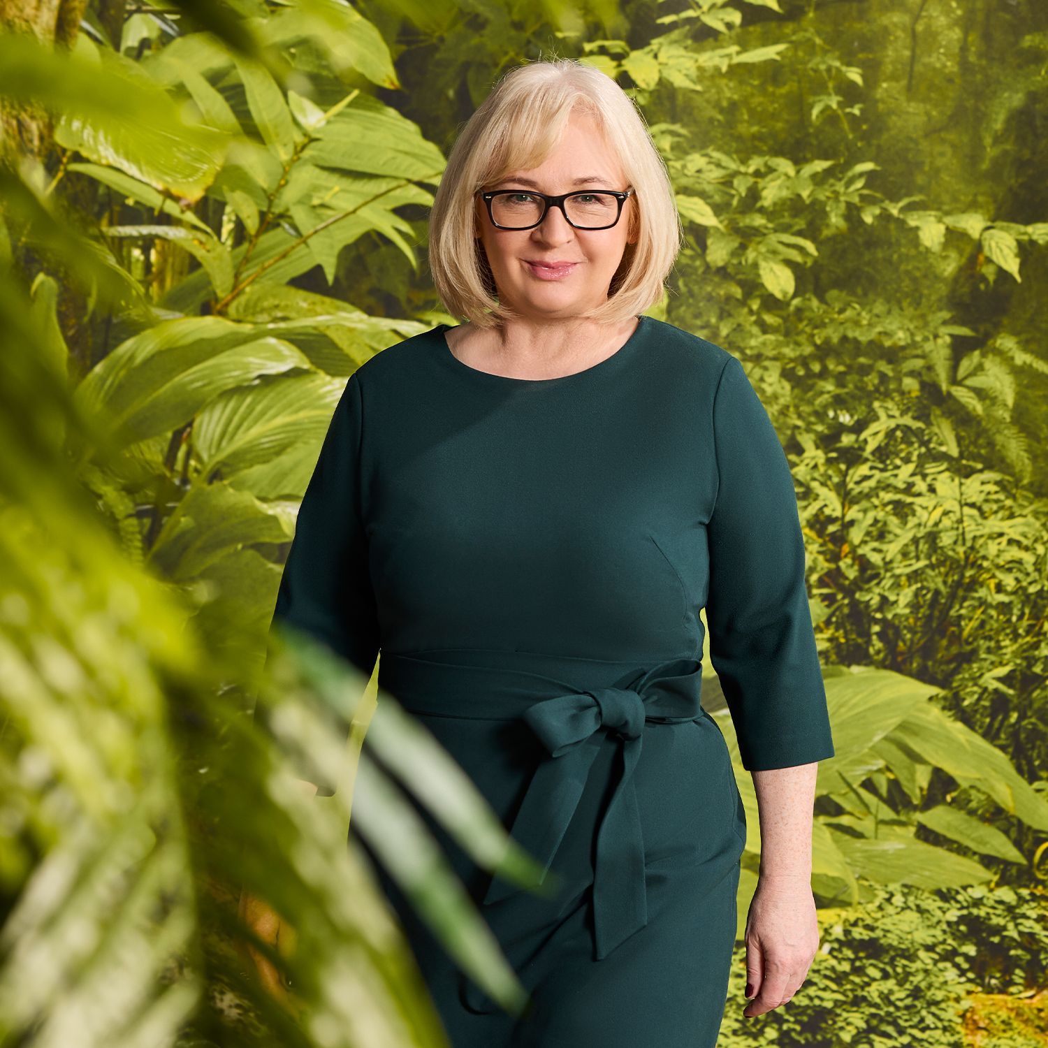 Urbrunnen-Expertin Monika Stedler trägt ein dunkelgrünes, knielanges Kleid mit 3/4-Ärmeln und einer Schleife an der Taille, steht vor dichtem grünem Laub