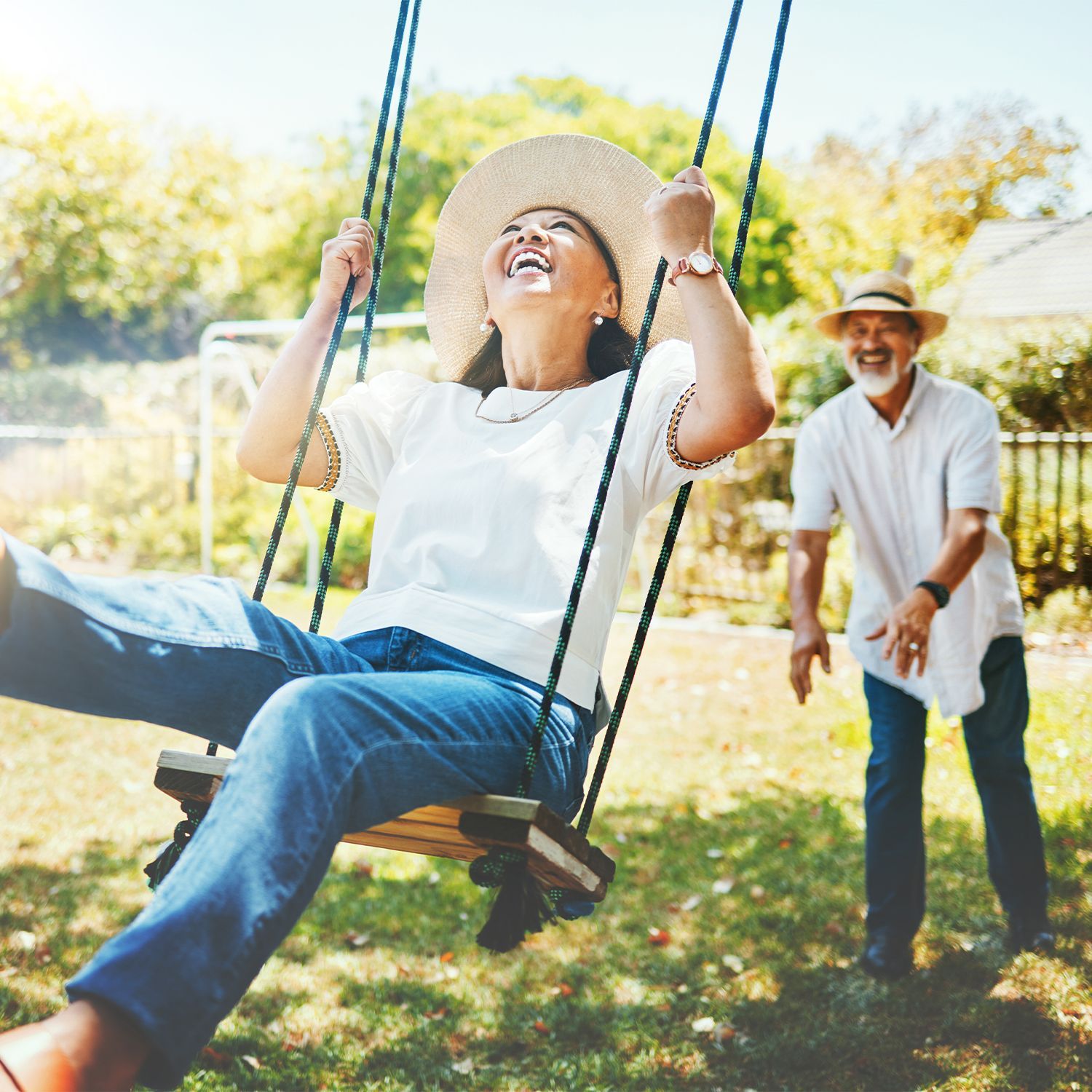 Ältere Frau mit Sonnenhut sitzt lachend auf einer Schaukel, ein älterer Mann mit Sonnenhund schubst sie lachend an.