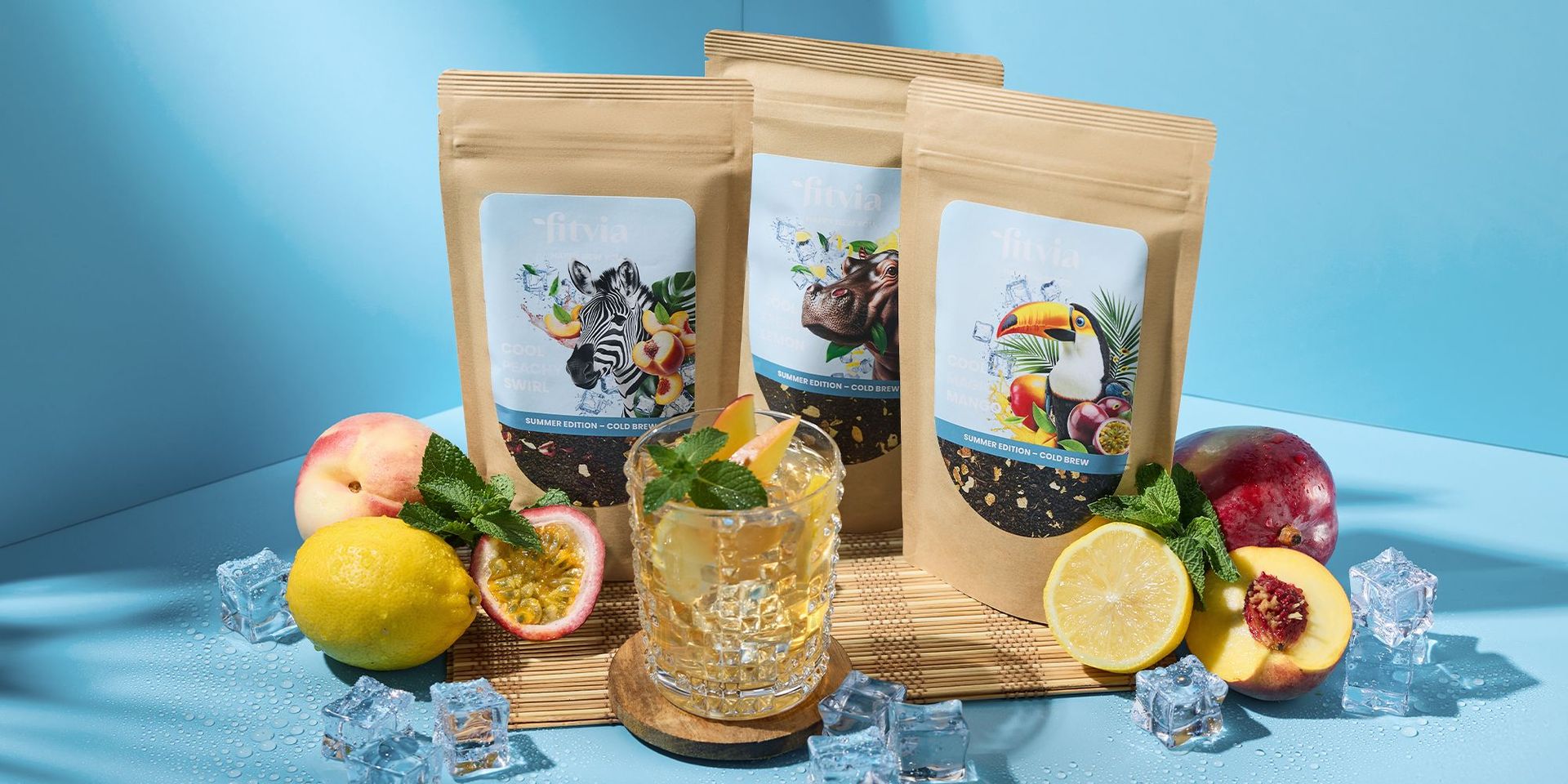 Drei braune Verpackungen mit Fitvia Cold Brew Tee, umgeben von Zitrone, Pfirsich, Passionsfrucht und Eiswürfeln, Glas mit Getränk und Minzblatt