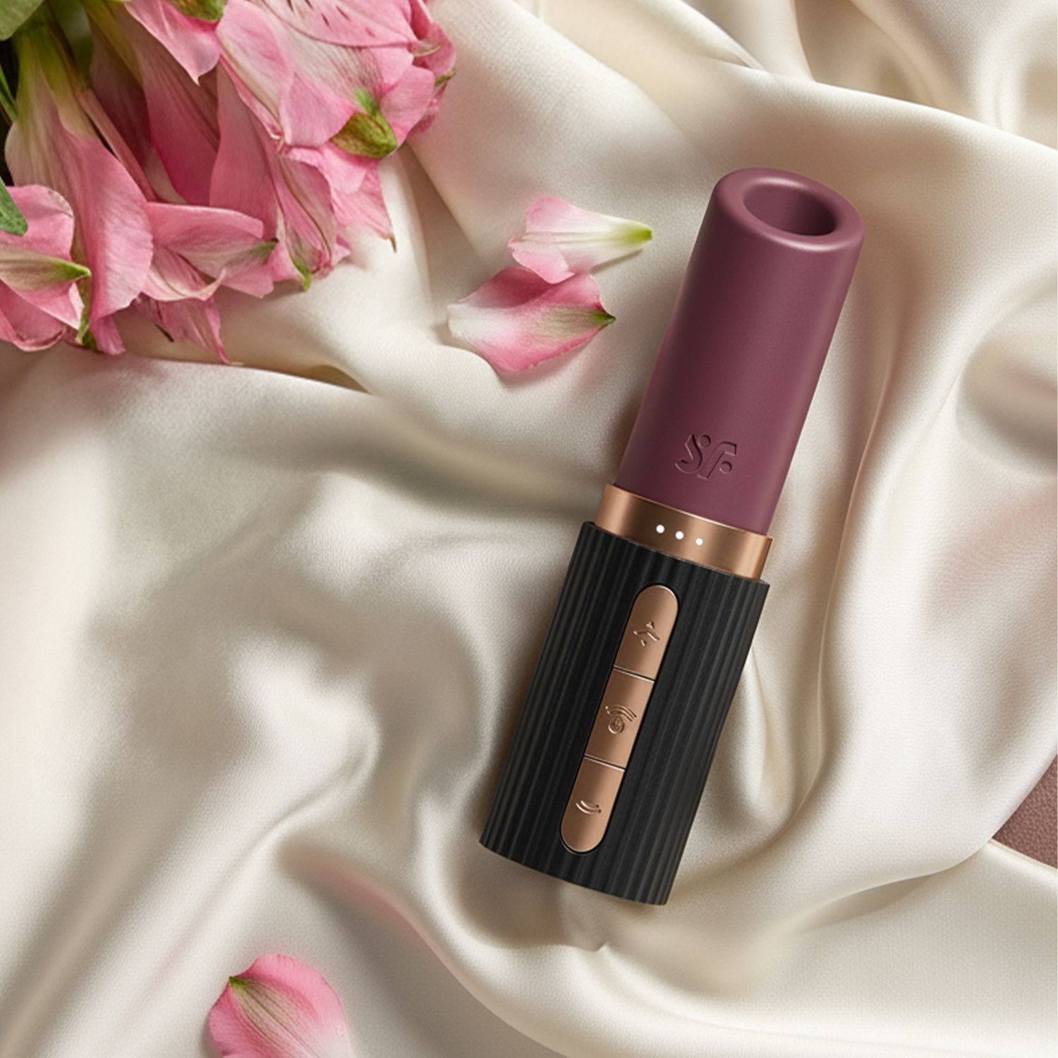 Satisfyer Massagegerät "Deep Kiss" im diskreten Lippenstift-Design auf weißem Satin Stoff und pinken Blüten