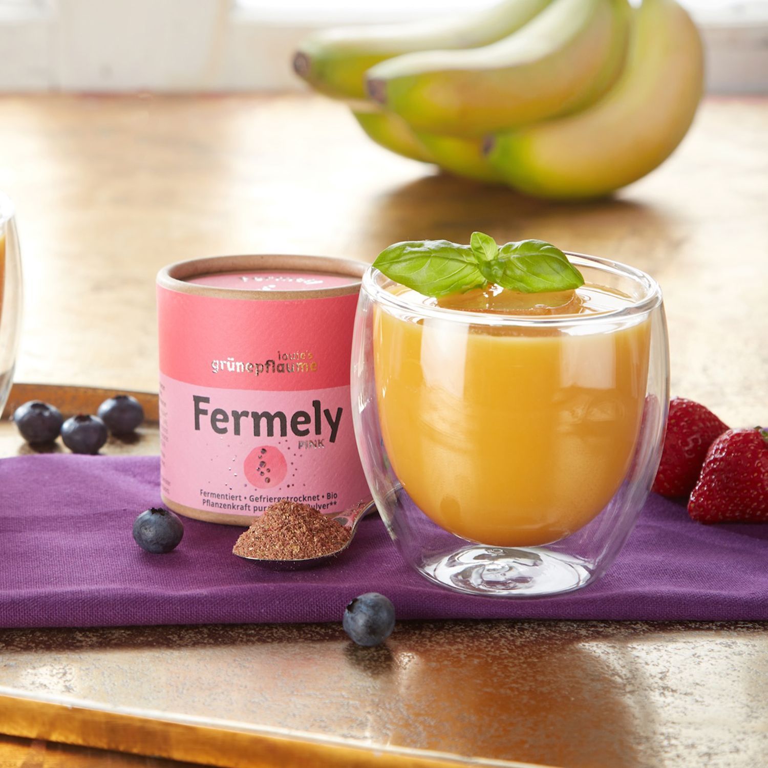 Zwei Gläser mit orangem Smoothie, jeweils mit Basilikumblatt, auf lila Tuch, daneben Dose mit Aufschrift 'Fermely pink' und verstreute Heidelbeeren, im Hintergrund Bananen und Erdbeeren.