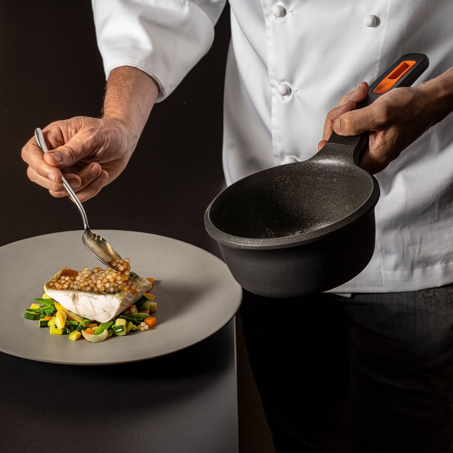 Hände eines Kochs, der mit einem Löffel Soße auf ein Fischfilet auf einem Gemüsebett auf einem grauen Teller gibt, in der anderen Hand eine schwarze Pfanne haltend.