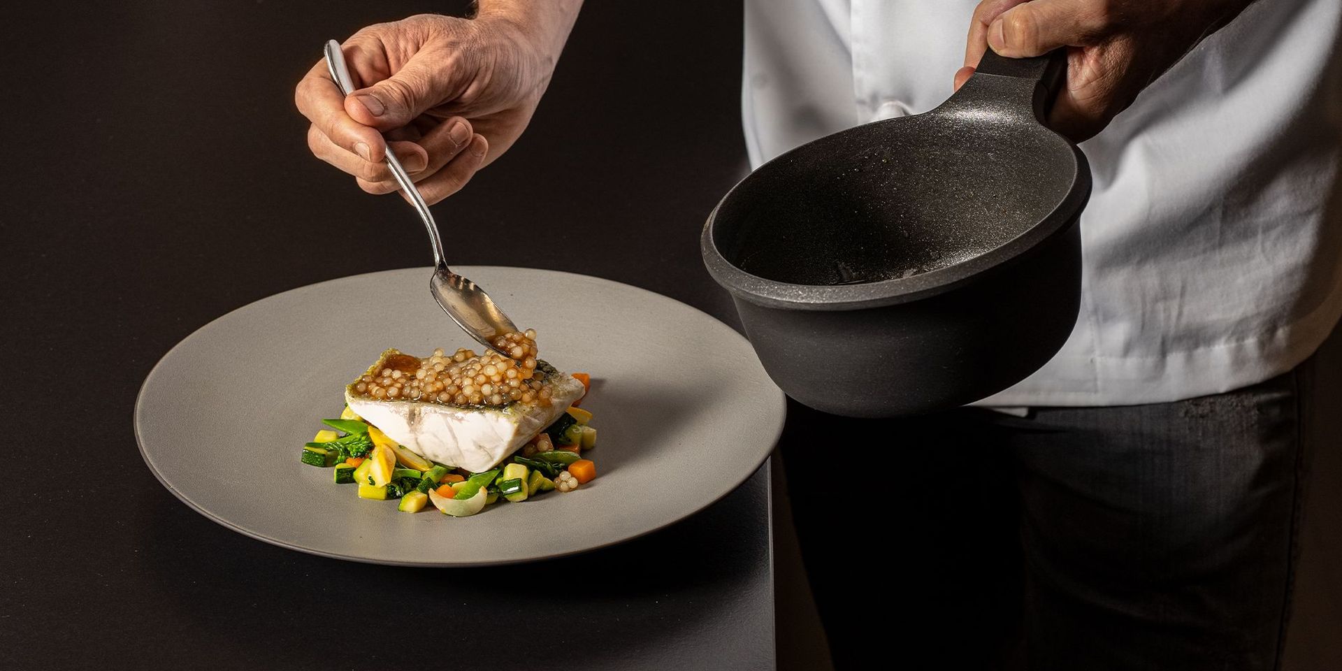 Hände eines Kochs, der mit einem Löffel Soße auf ein Fischfilet auf einem Gemüsebett auf einem grauen Teller gibt, in der anderen Hand eine schwarze Pfanne haltend.