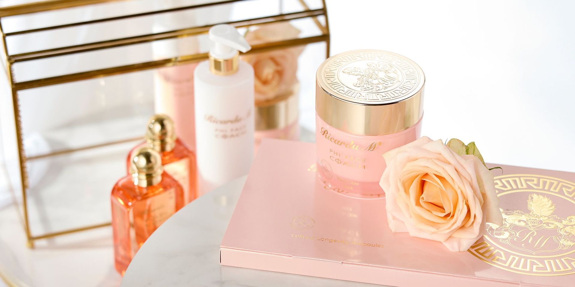 Pfirsichfarbene Kosmetikprodukte von Ricarda M., darunter ein Gesichtscreme-Tiegel mit goldfarbenem Deckel, eine Augencreme mit goldfarbenen Enden und eine Flasche, neben einer hellrosa Rose auf einem weißen Tisch.