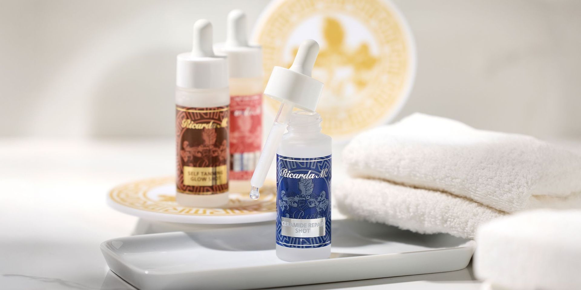 Drei Ricarda M Hautpflege-Shots in Flaschen mit Pipetten, vorn Ceramide Repair Shot in blauer Flasche, dahinter Self Tanner Glow Shot und ein weiterer Shot, daneben weiße Handtücher und gold-weiße Untersetzer.