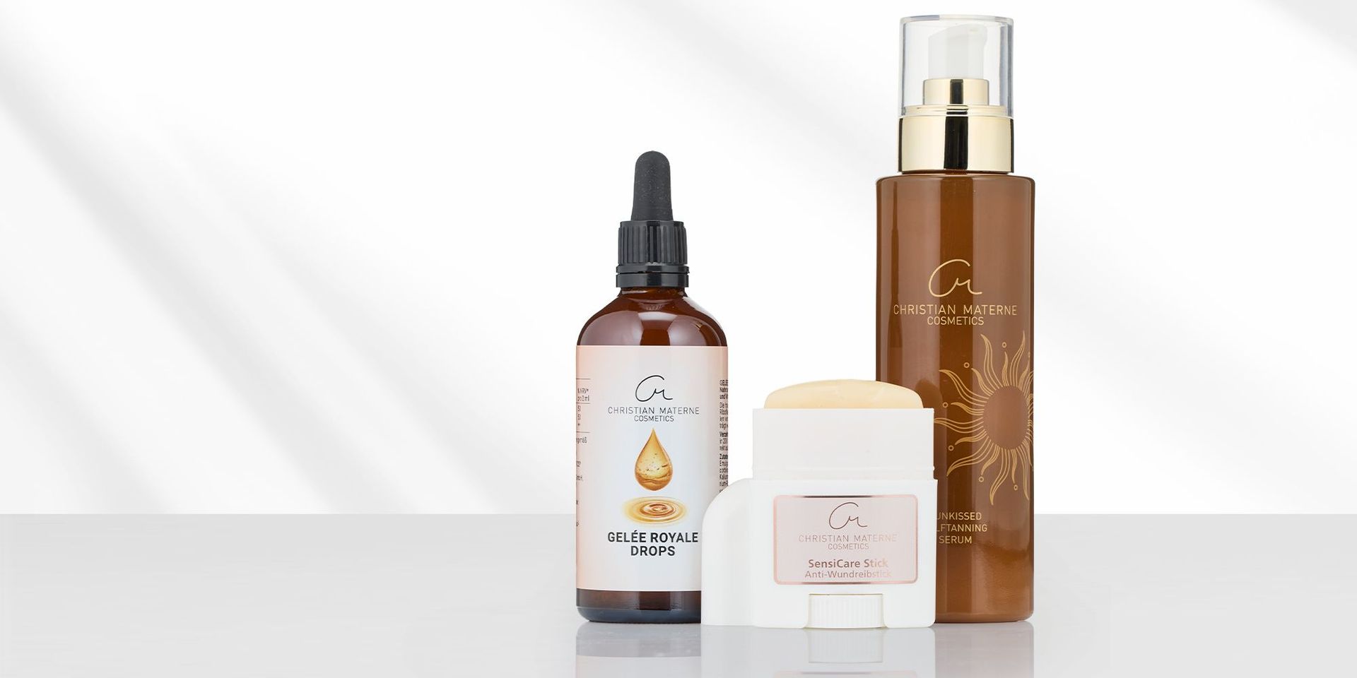 Drei Kosmetikprodukte von Christian Materne Cosmetics: braune Flasche mit Pipette, weißer Stick mit Deckel, braune Flasche mit Pumpspender und Sonnensymbol.