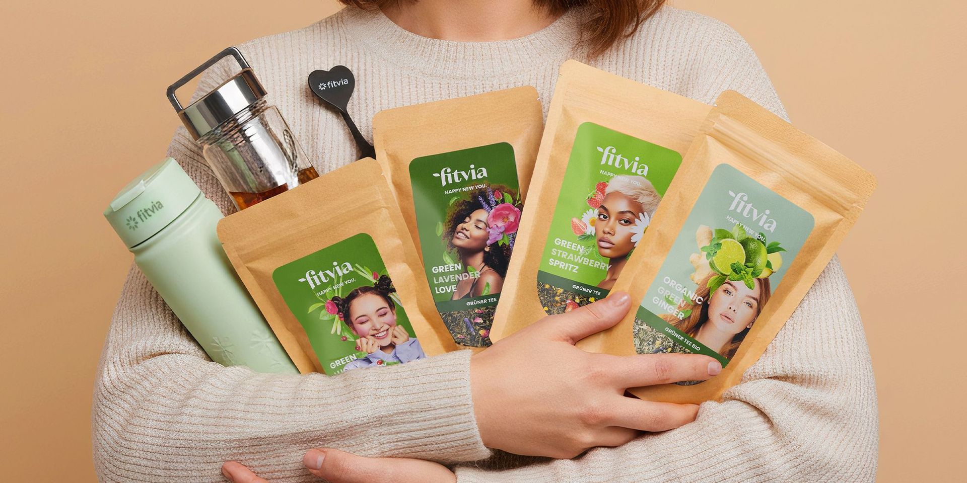 Frau in beigem Pullover hält mehrere braune Papiertüten mit grünen fitvia-Etiketten und eine grüne fitvia-Trinkflasche sowie einen Teefilter mit Griff.