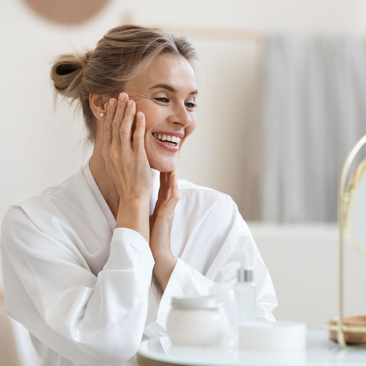Frau im weißen Bademantel berührt ihr Gesicht vor einem runden Spiegel auf einem Tisch mit Kosmetikbehältern.