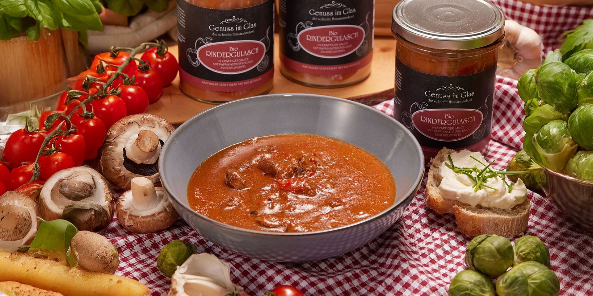Graue Schale mit braunem Bio-Rindergulasch, umgeben von Pilzen, Tomaten, Brot und Gläsern mit Bio-Rindergulasch-Sauce.