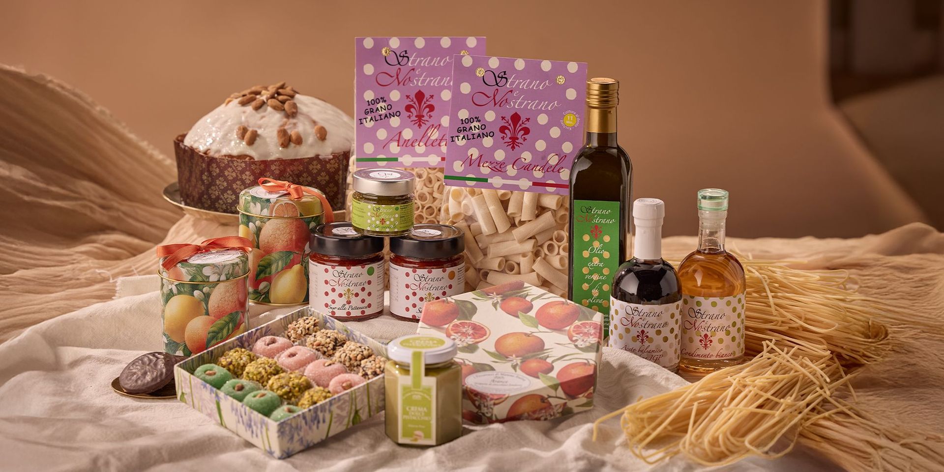 Verschiedene italienische Lebensmittel: Panettone mit Zuckerguss und Sonnenblumenkernen, Gläser mit Marmelade und Pesto, bunte Kekse, zwei Packungen Pasta, Flaschen mit Öl und Essig, lose Spaghetti auf Stoff.