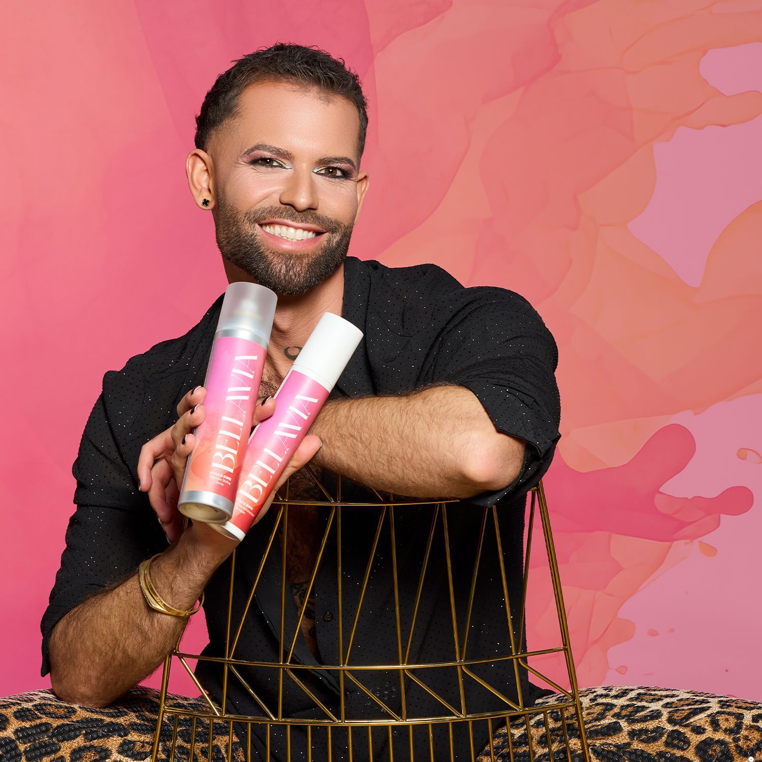 Person mit schwarzem Hemd und Leopardenhose hält zwei pink-weiße Bellavia-Sprühflaschen vor einem rosa Hintergrund.