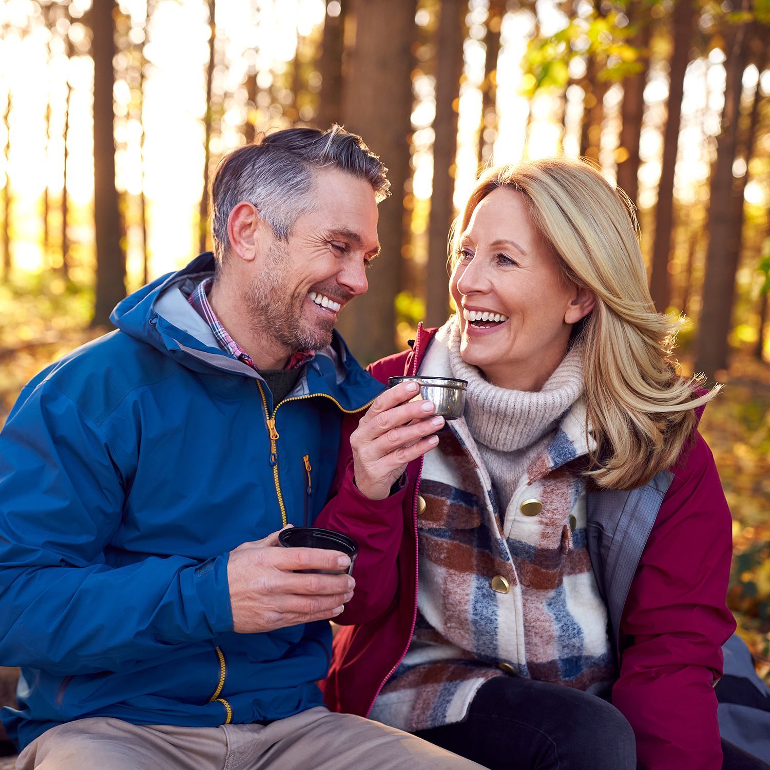 Mann und Frau im Wald, beide tragen Jacken, der Mann hält einen Becher, die Frau hält eine Thermoskanne mit Ausgießer.