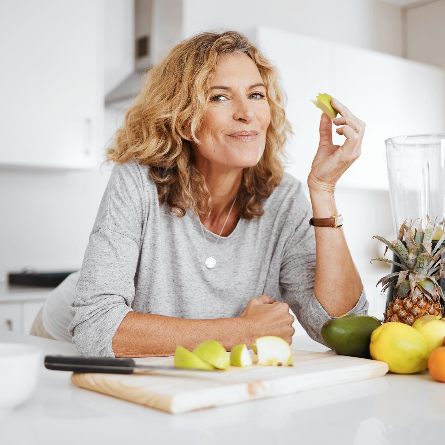 Frau mit grauem Pullover sitzt am Küchentisch, hält ein Stück Obst in der Hand, auf dem Tisch liegen verschiedene Früchte und ein Mixer.