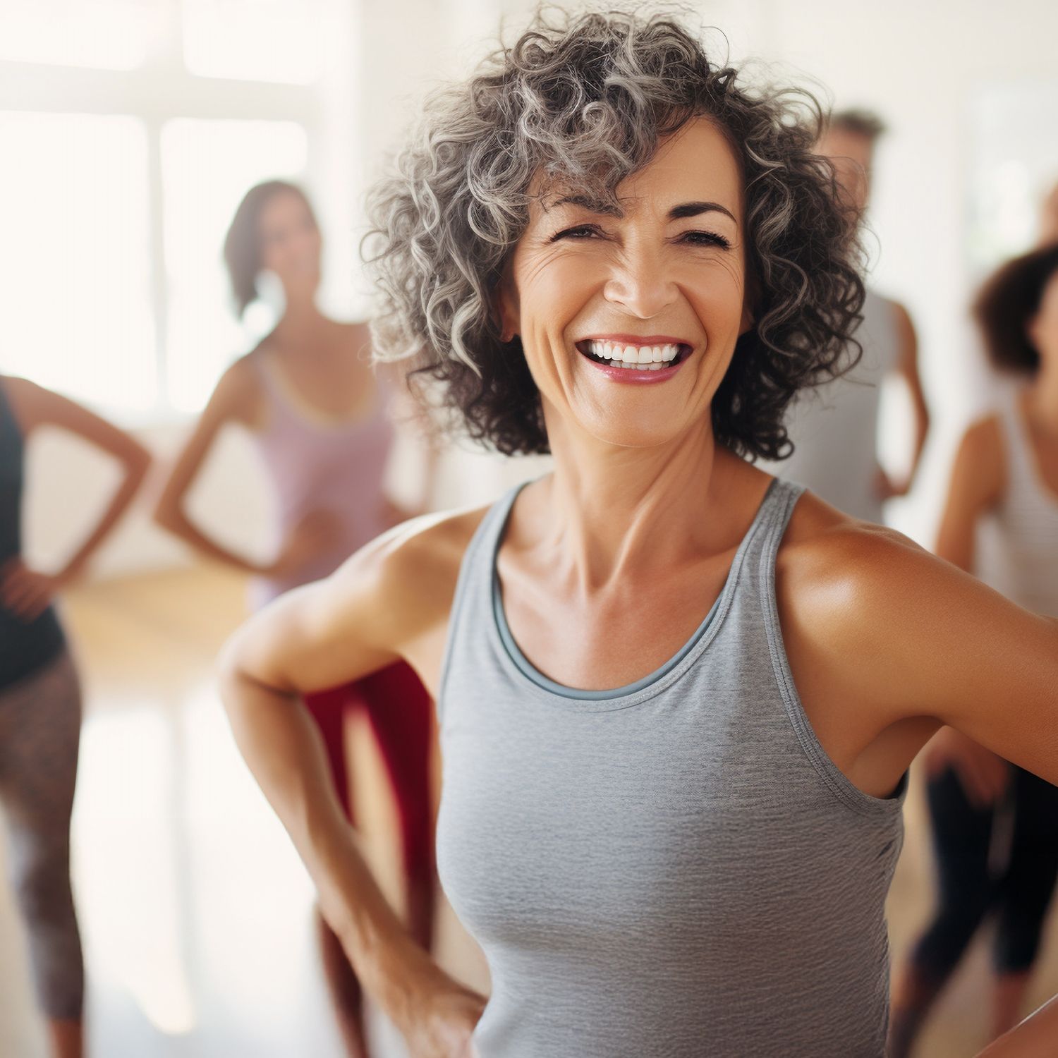 Mehrere Personen in Sportkleidung stehen mit Händen in der Hüfte in einem hellen Raum, vorn eine Frau mit lockigem Haar und grauem Tanktop.
