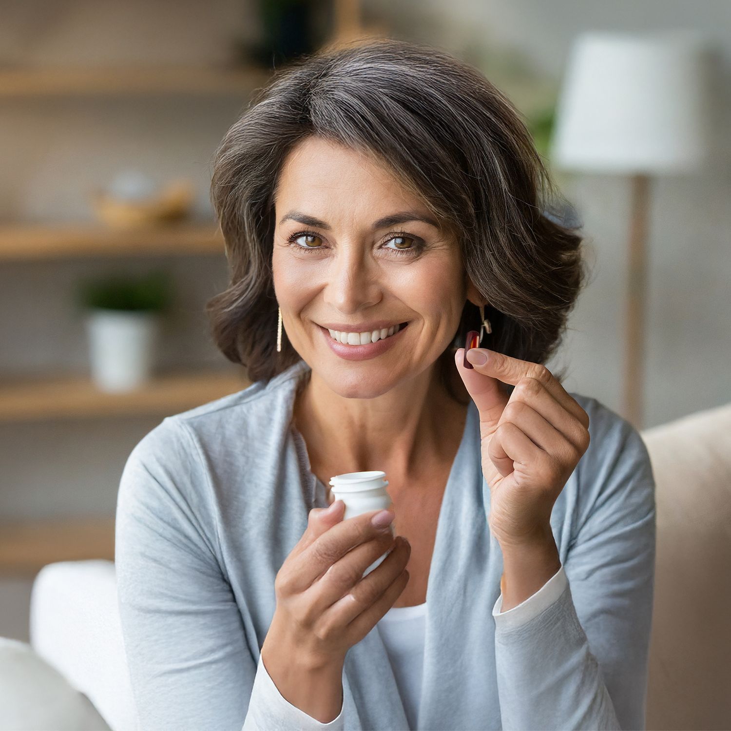 Frau mit grauem Pullover hält eine weiße Tablettenflasche in der linken Hand und eine Tablette in der rechten Hand, im Hintergrund ein Regal mit Pflanzen und Lampen.