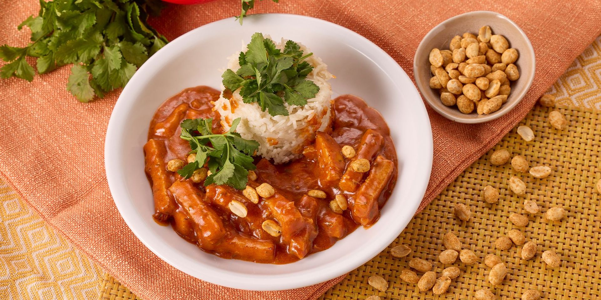 Weißer Teller mit Hähnchencurry in roter Sauce, garniert mit Erdnüssen und Koriander, dazu eine Portion Reis mit Korianderzweig.