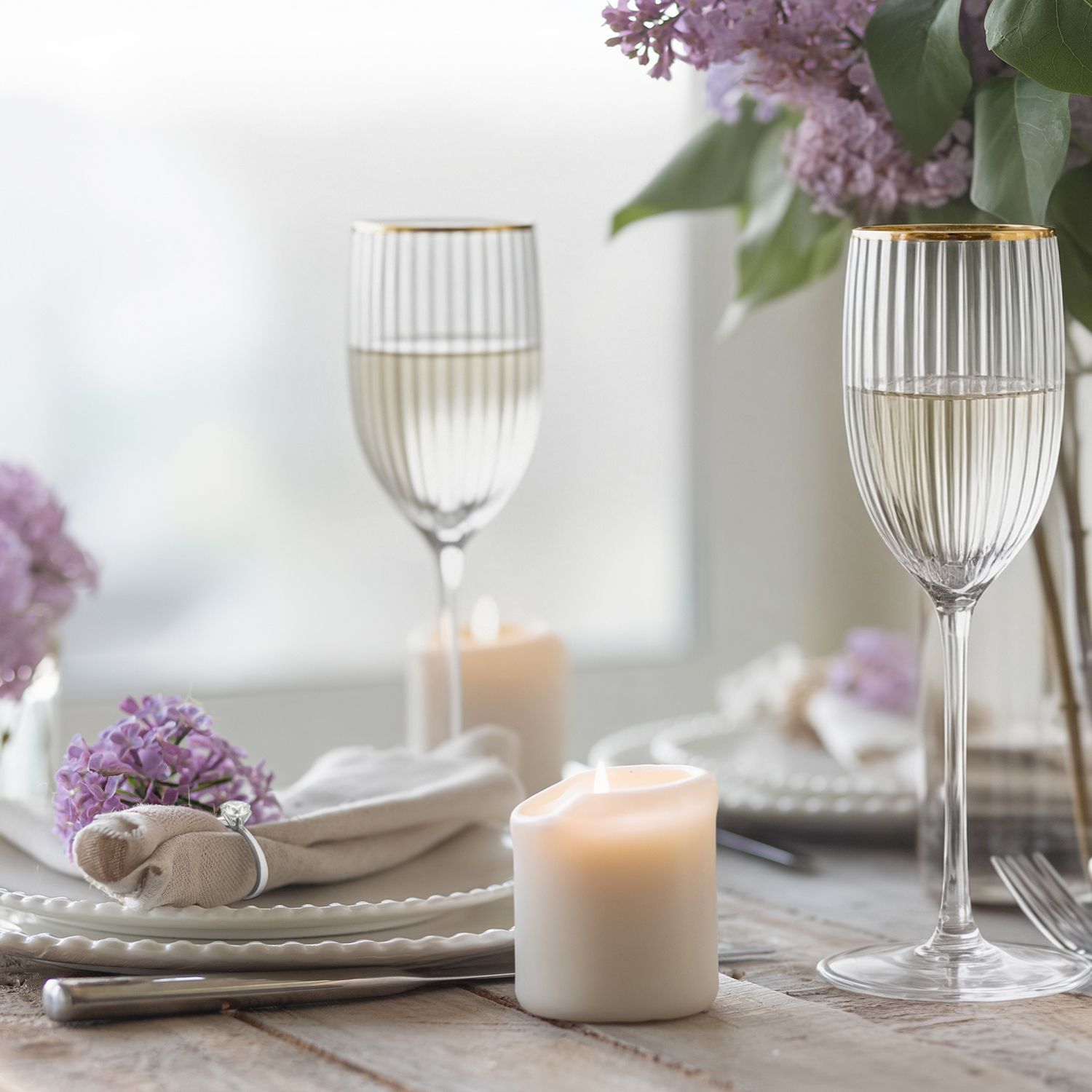 Gedeckter Holztisch mit weißen Kerzen, goldrandigen Weingläsern, weißen Tellern, Stoffservietten und lila Fliederblüten