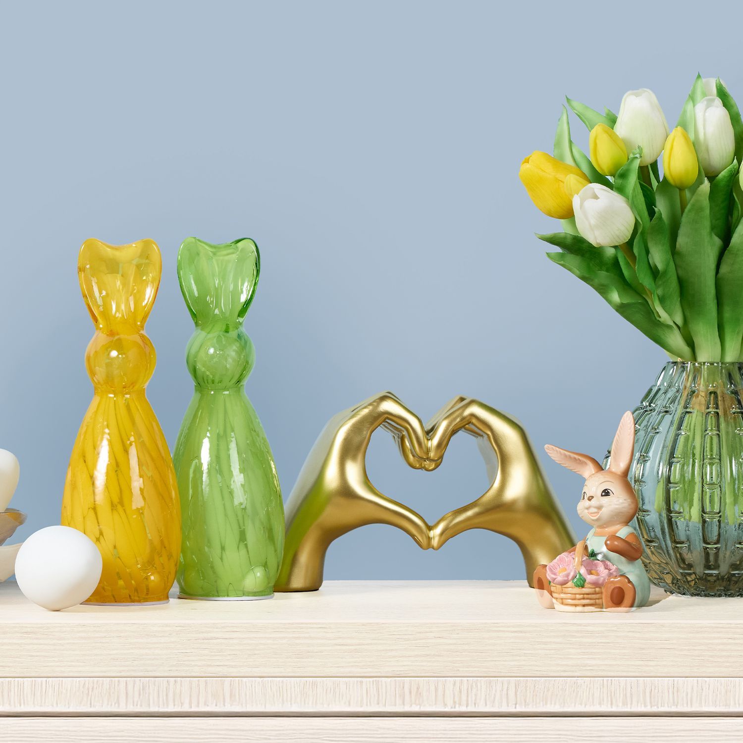 Gelbe und grüne Hasenfiguren aus Glas, goldene Hände, die ein Herz formen, weiße Eier in Schalen und ein Glasvase mit gelben und weißen Tulpen vor blauem Hintergrund