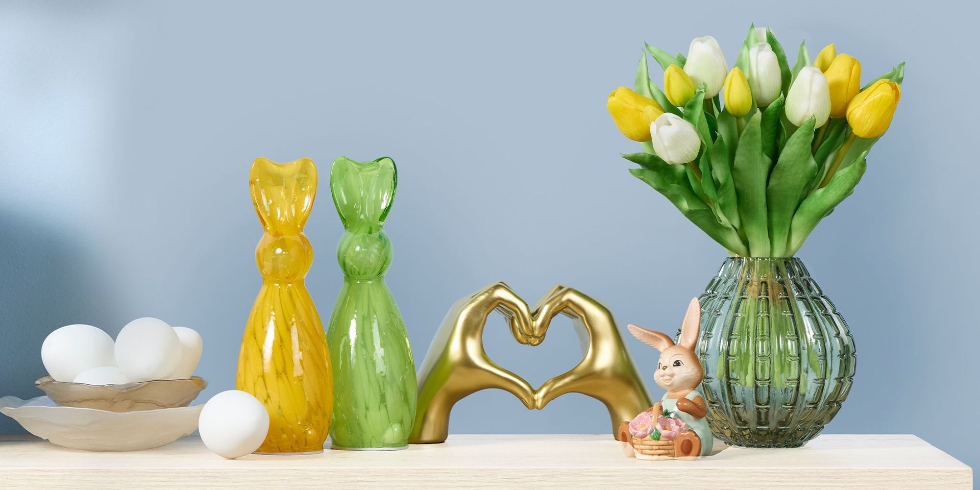 Gelbe und grüne Hasenfiguren aus Glas, goldene Hände, die ein Herz formen, weiße Eier in Schalen und ein Glasvase mit gelben und weißen Tulpen vor blauem Hintergrund