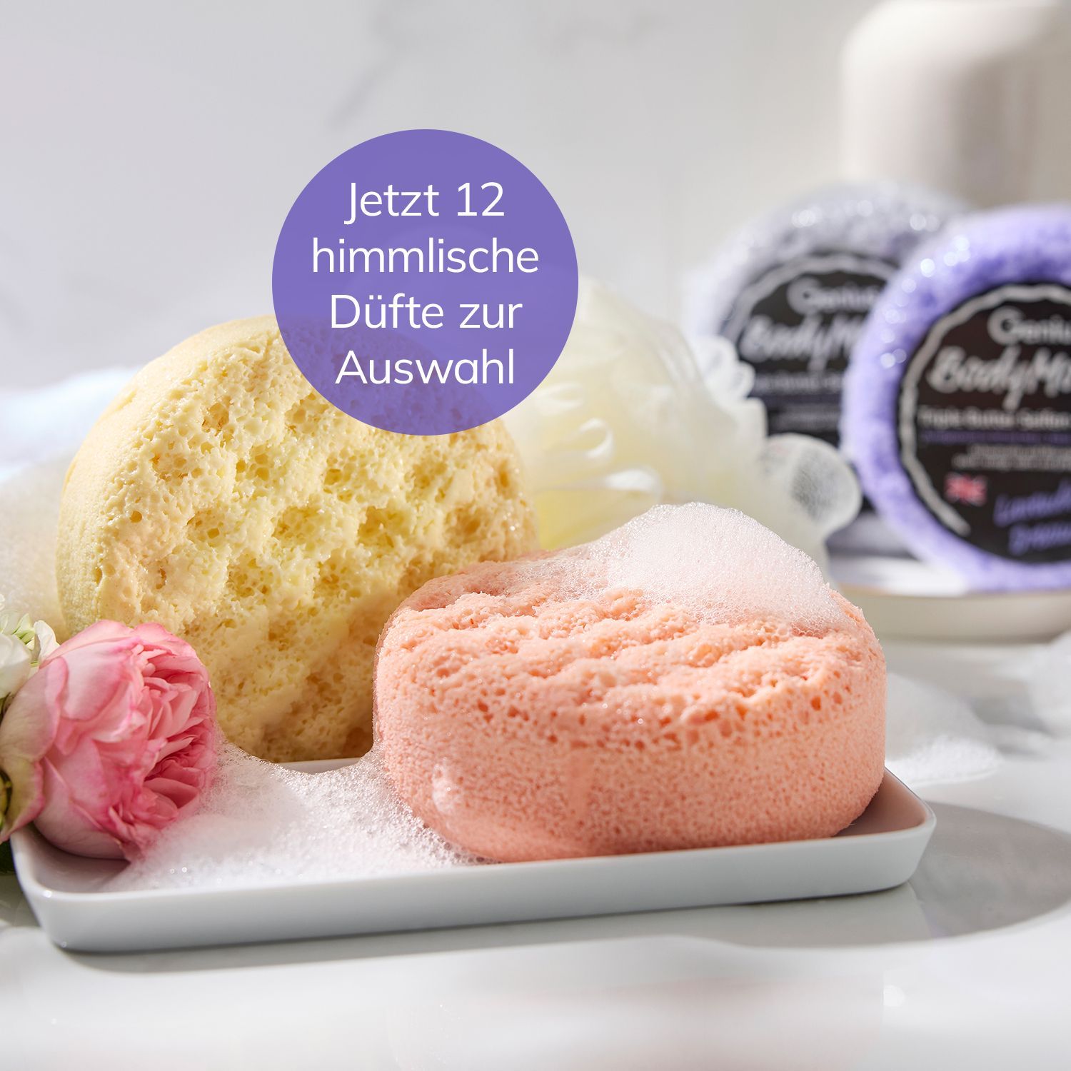 Zwei Körperpflegeschwämme auf weißem Teller mit rosa Blume und Verpackungen im Hintergrund.