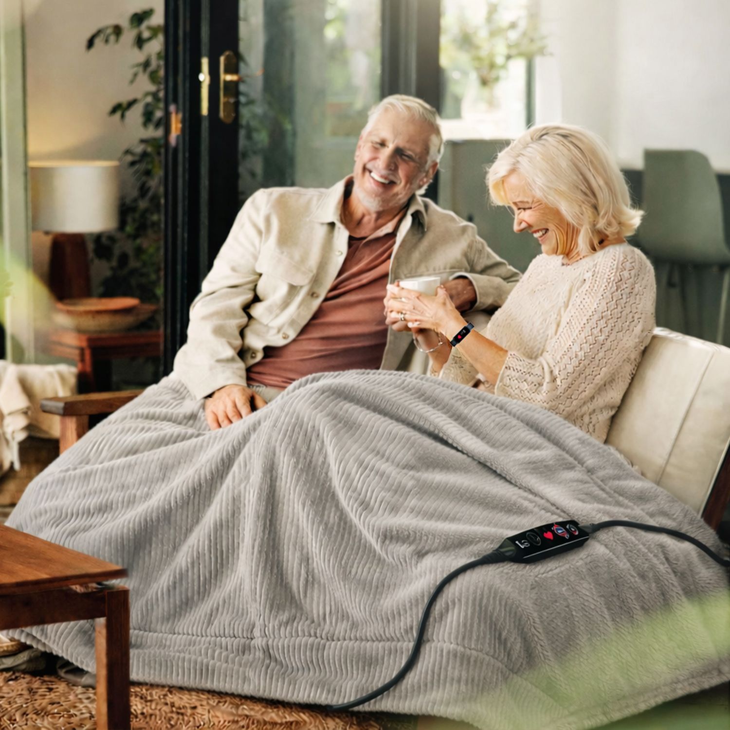 Älteres Paar sitzt auf Sofa unter einer grauen, gerippten Heizdecke mit Fernbedienung