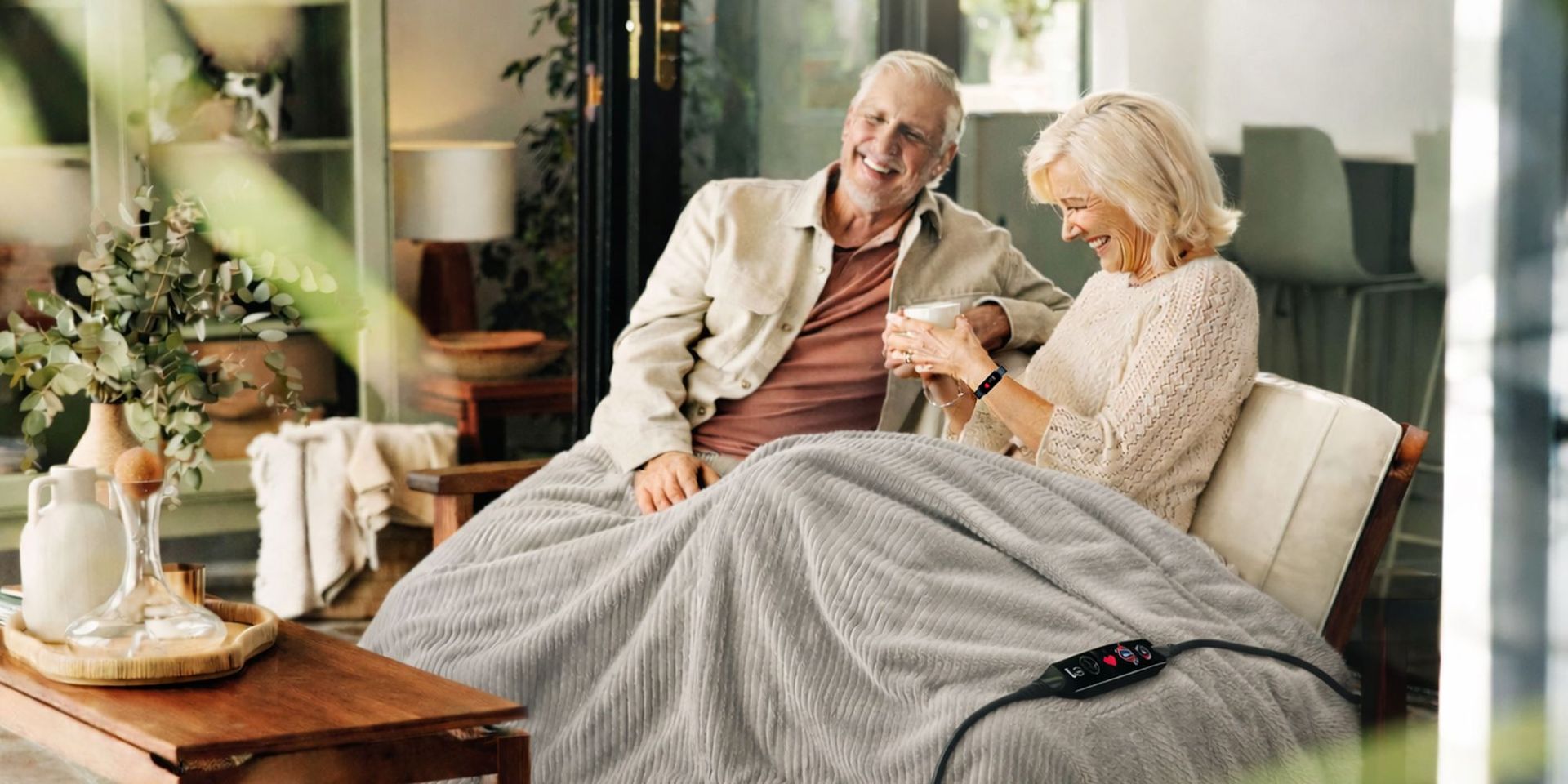 Älteres Paar sitzt auf Sofa unter einer grauen, gerippten Heizdecke mit Fernbedienung