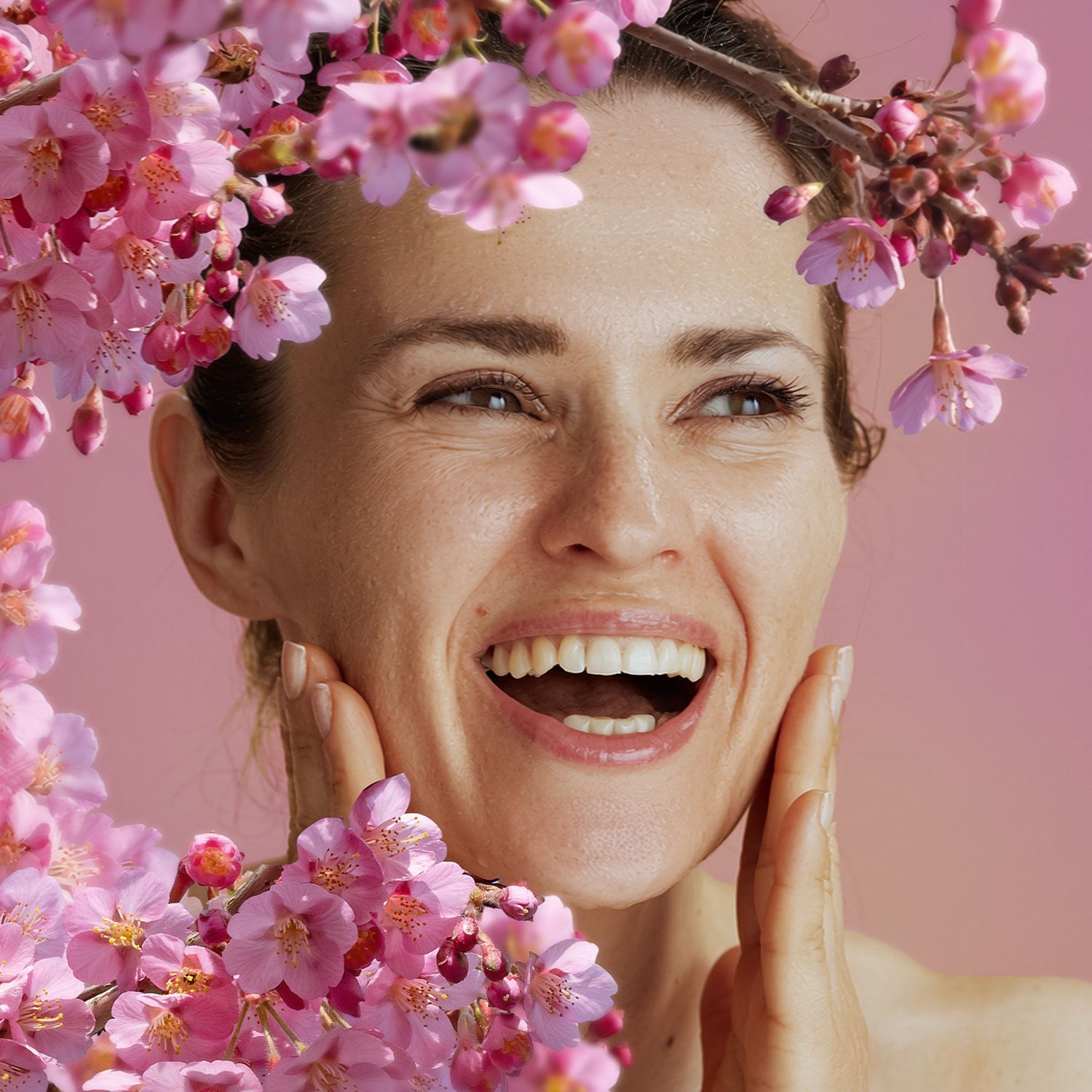 Dunkelhaarige Frau, die ihre Hände ans Gesicht hält und teilweise sichtbare Schulter vor rosa Hintergrund, davor Zweige mit zahlreichen kleinen Blüten  