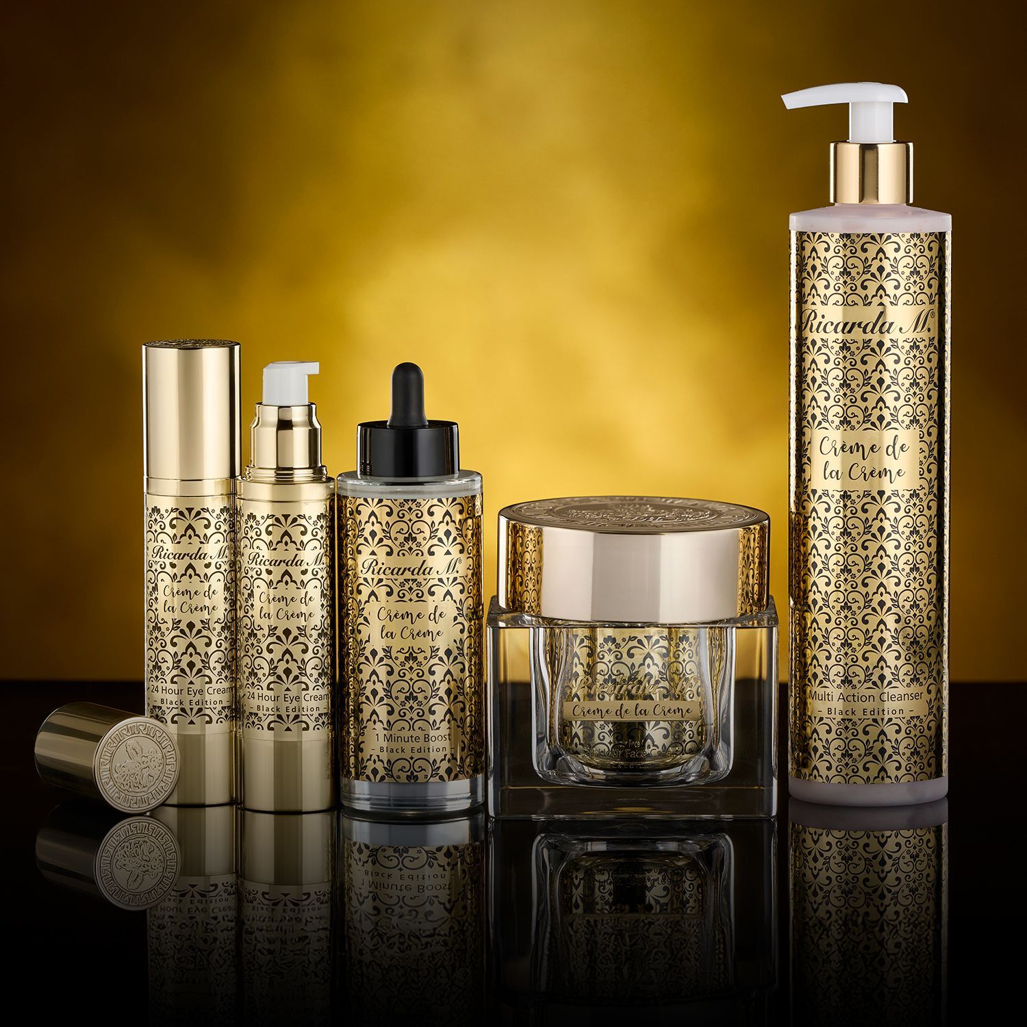 Fünf goldfarbene Kosmetikbehälter mit schwarzem Ornamentmuster und Pump- oder Tropfverschluss auf schwarzem Untergrund vor goldfarbenem Hintergrund