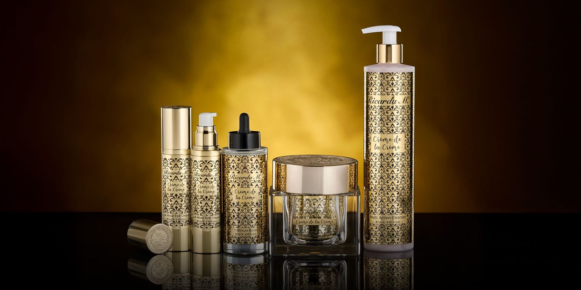 Fünf goldfarbene Kosmetikbehälter mit schwarzem Ornamentmuster und Pump- oder Tropfverschluss auf schwarzem Untergrund vor goldfarbenem Hintergrund