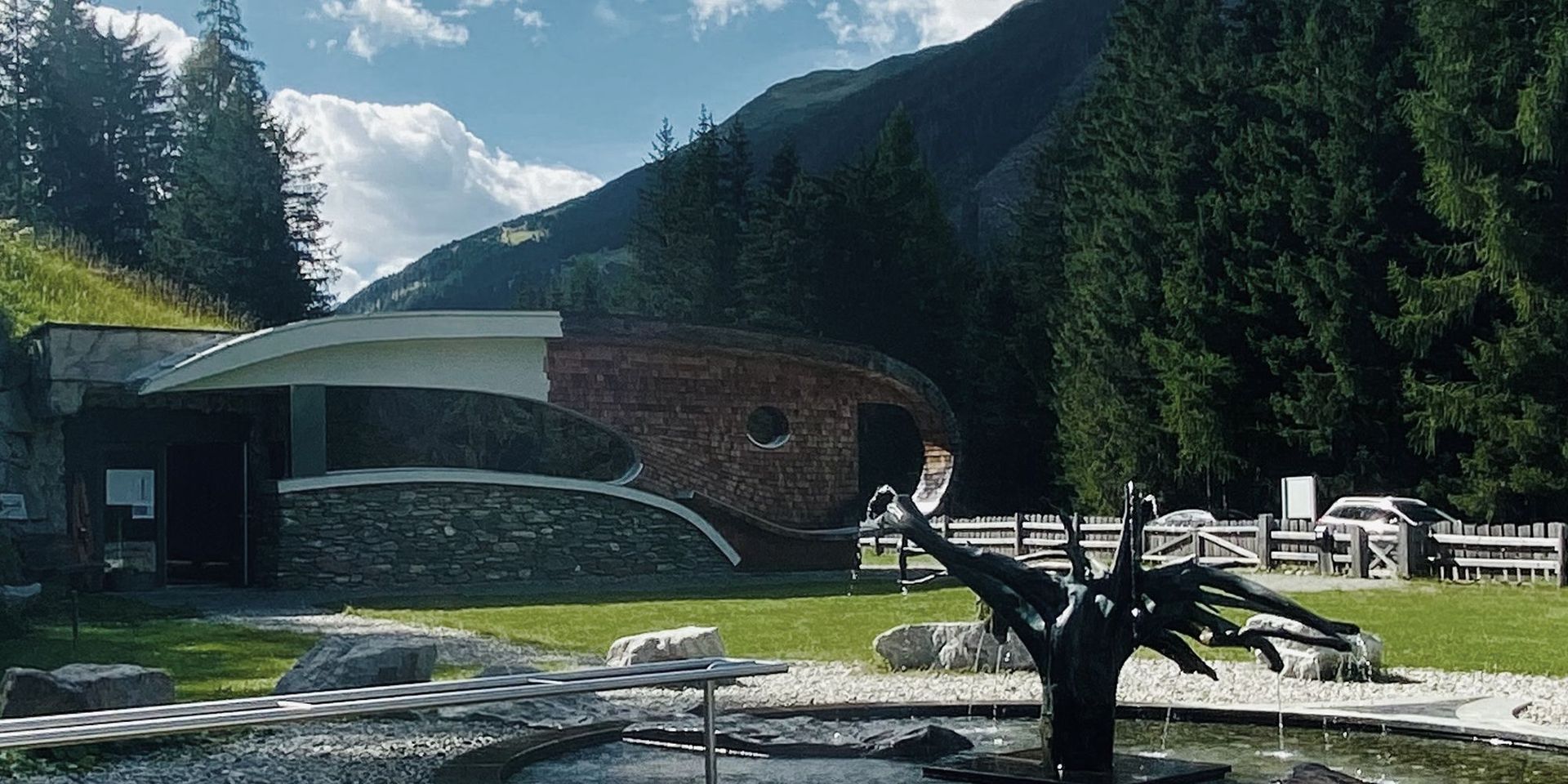 Deferegger Heilwasser – die Quelle Moderne Architektur in alpiner Landschaft, umgeben von Bergen und Bäumen, im Kontext von Mobilität und Freizeit.