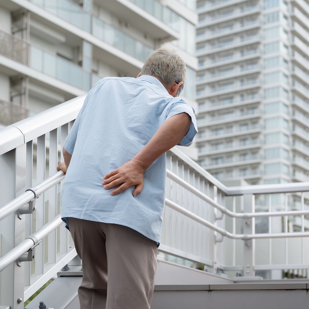 Rückenschmerzen Älterer Mann stützt sich mit einer Hand auf ein Treppengeländer und hält sich mit der anderen Hand den unteren Rücken.