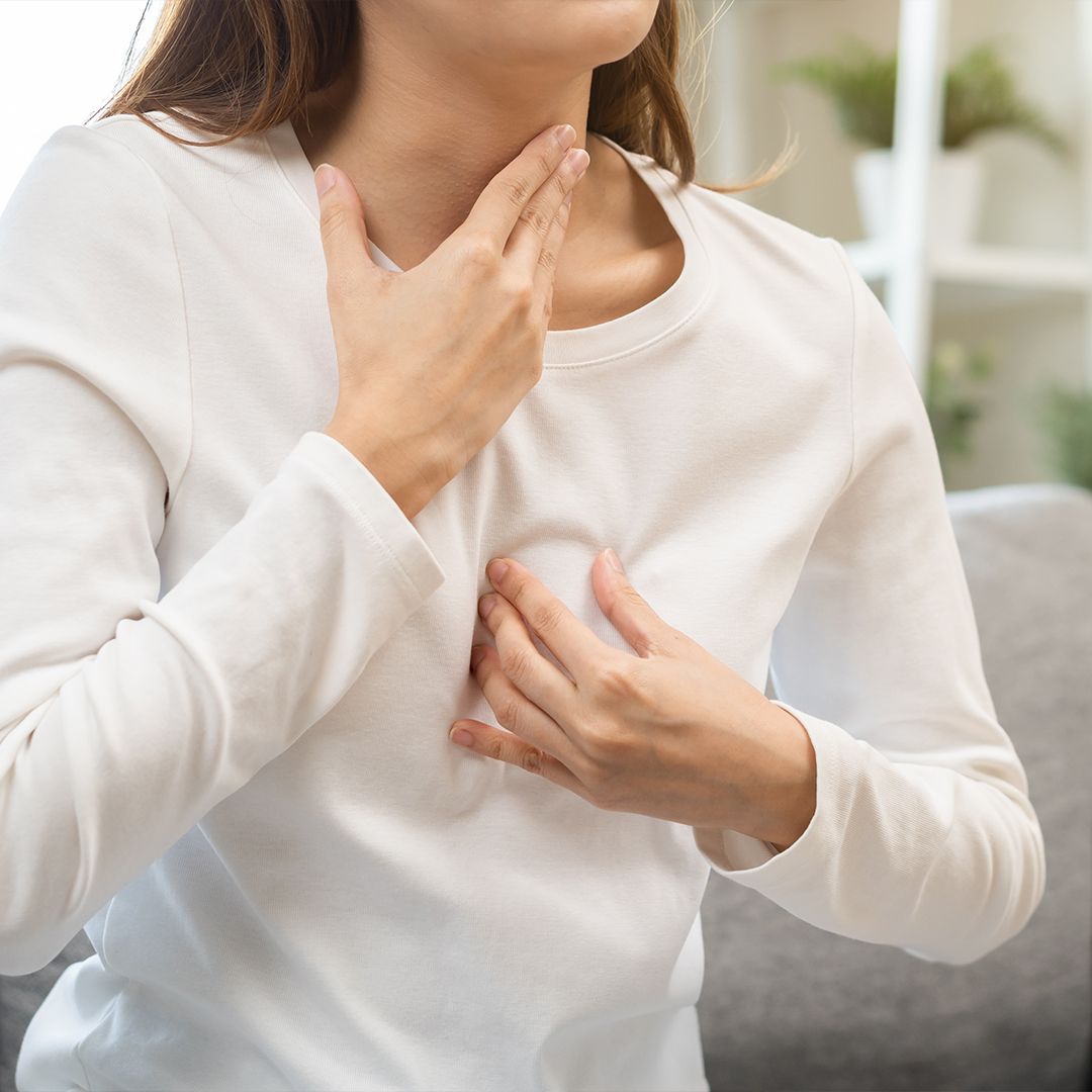 Sodbrennen Frau in weißem Langarmshirt sitzt auf grauem Sofa und hält eine Hand an die Brust, die andere an den Hals.
